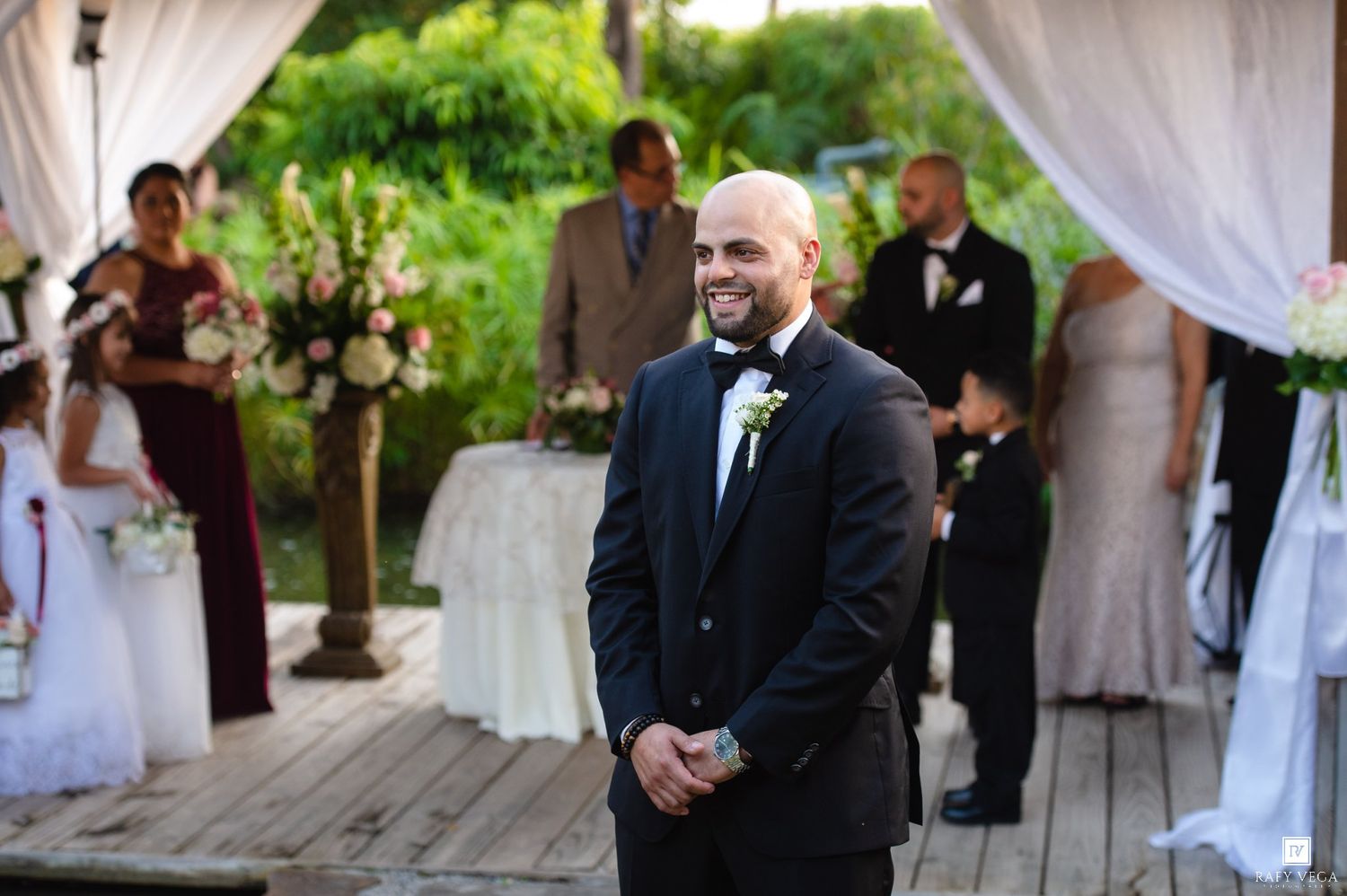 Groom smiling at outdoor wedding ceremony on wooden deck with bridal party and floral arrangements.