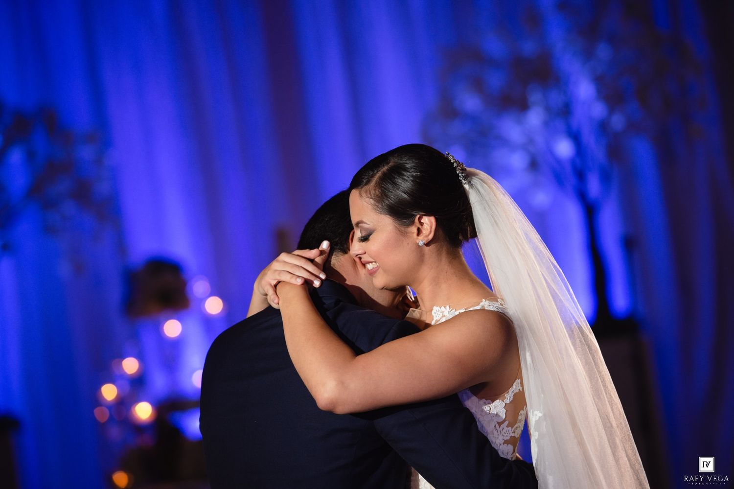Bride in white gown and veil shares emotional first dance embrace with groom under blue wedding reception lighting.