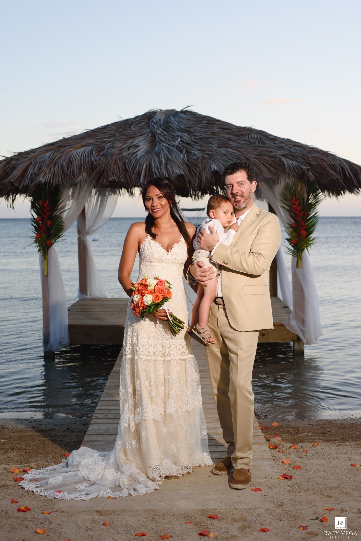 Bride and groom holding baby at beachside wedding ceremony under thatched gazebo with tropical decor.