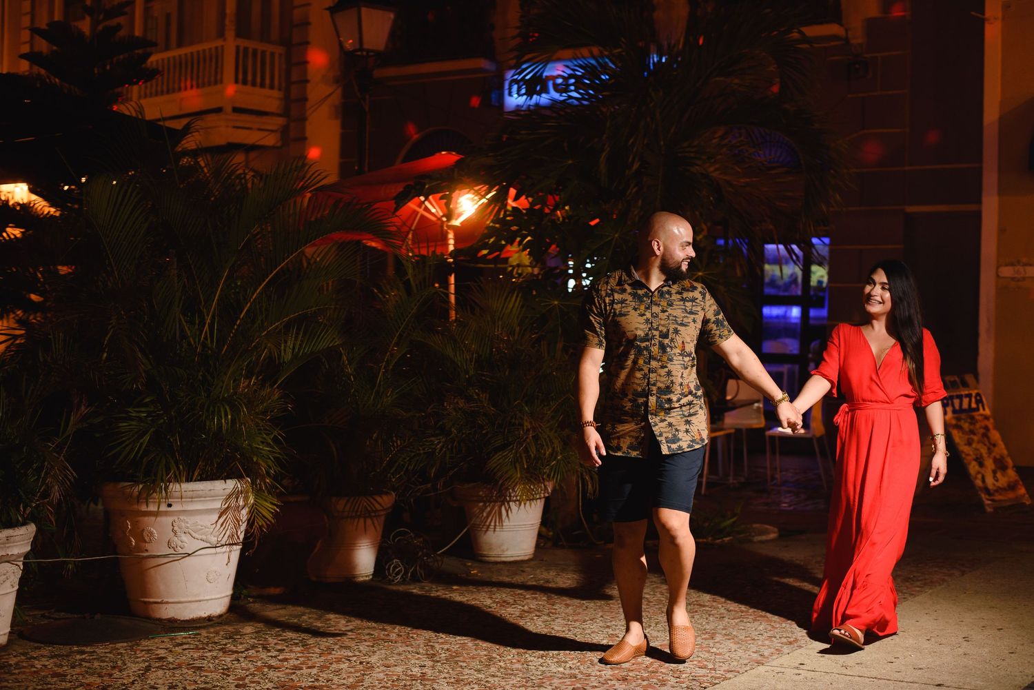 Couple holding hands walking at night past tropical plants and warm lights in a romantic outdoor setting.