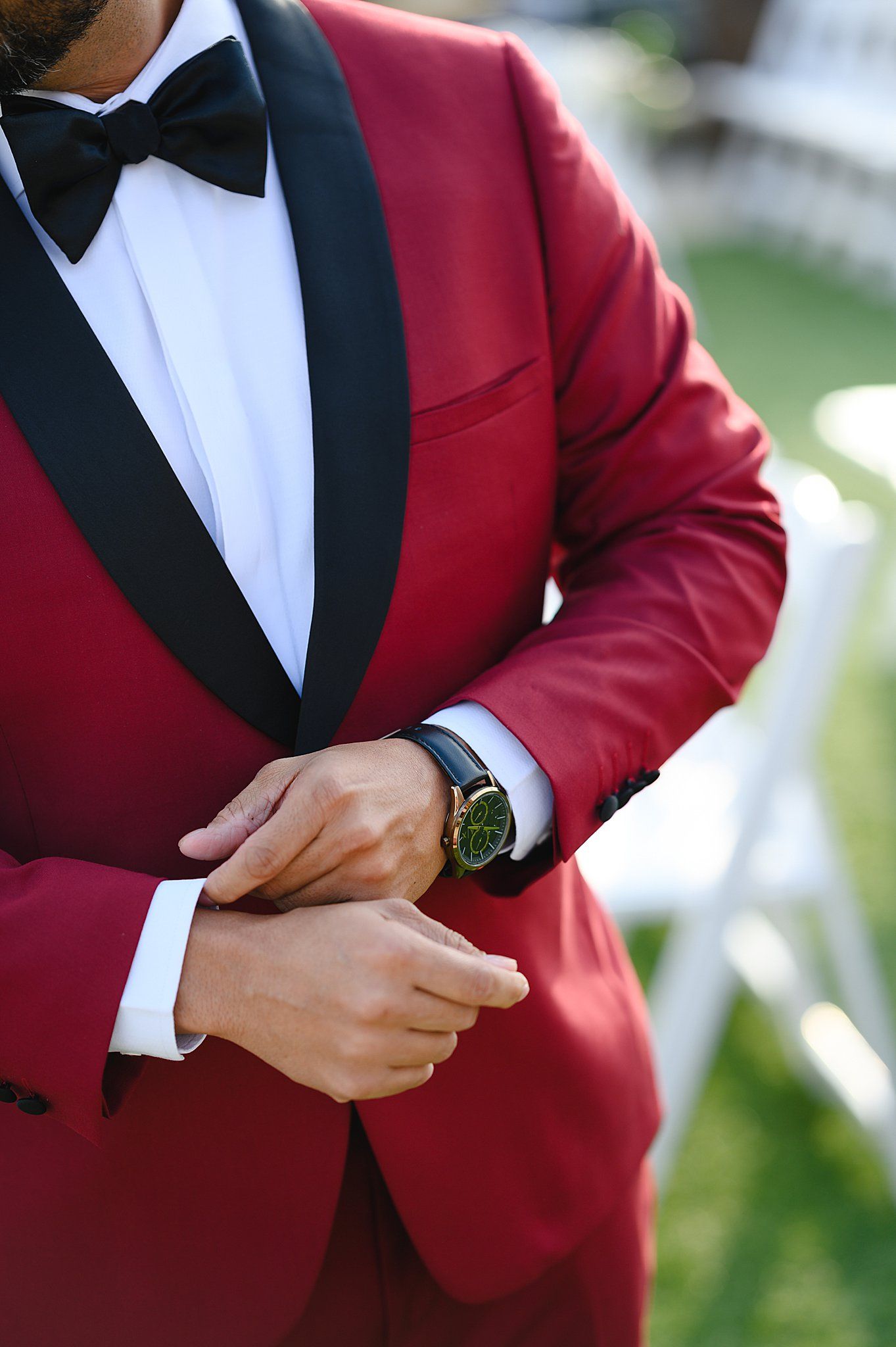 Man in red tuxedo with black lapels adjusting cufflinks, wearing a green watch and black bow tie at formal event.