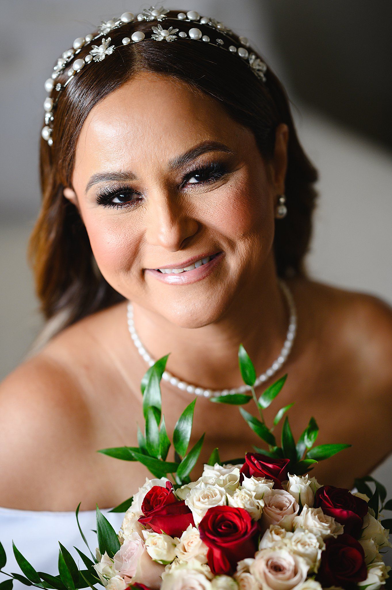 Smiling bride wearing pearl necklace and floral headband holds bouquet of red and white roses on her wedding day.