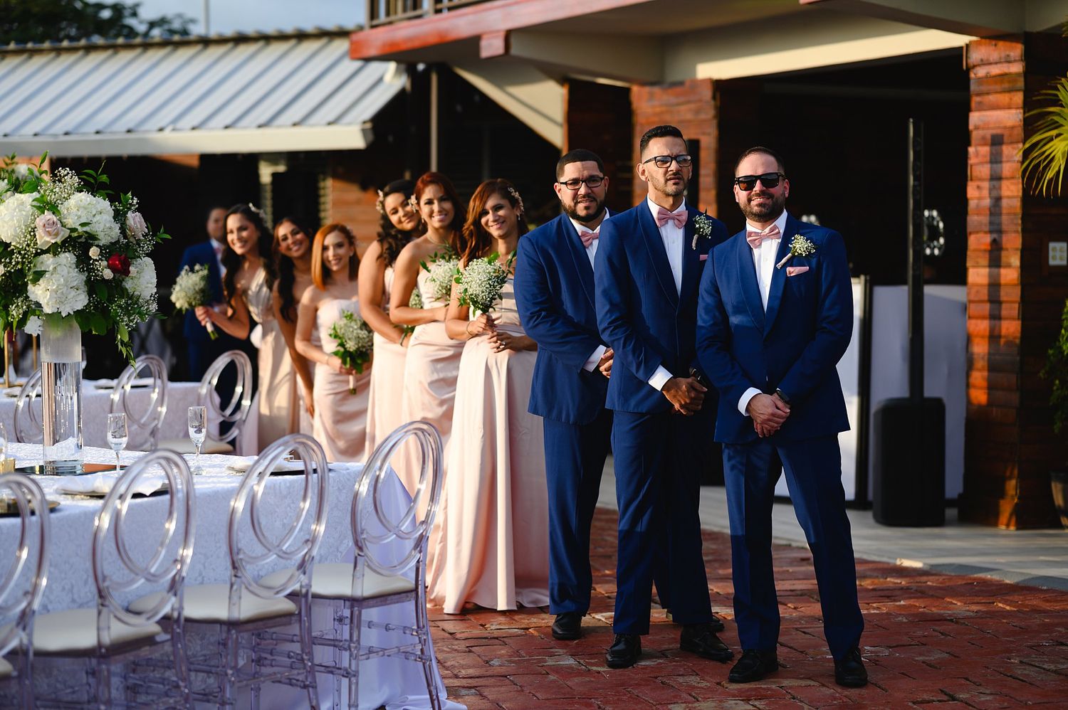 Groomsmen in navy suits stand with bridesmaids in blush gowns at an elegant outdoor wedding reception venue.