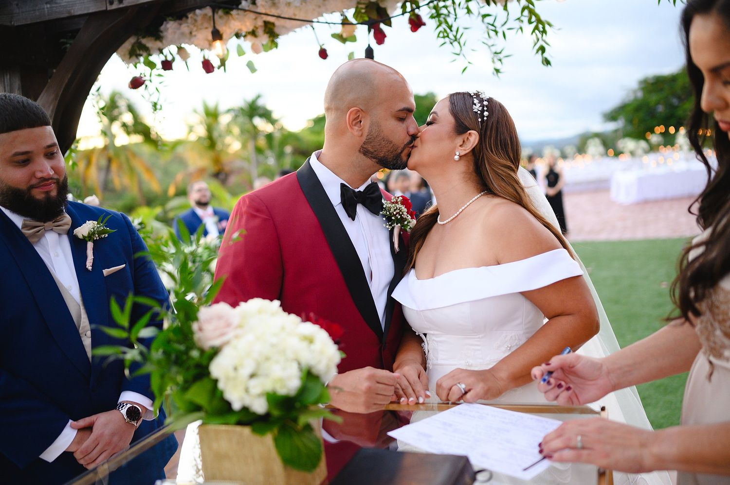 Bride and groom share first kiss at outdoor wedding ceremony, groom in red tuxedo, bride in off-shoulder white gown.