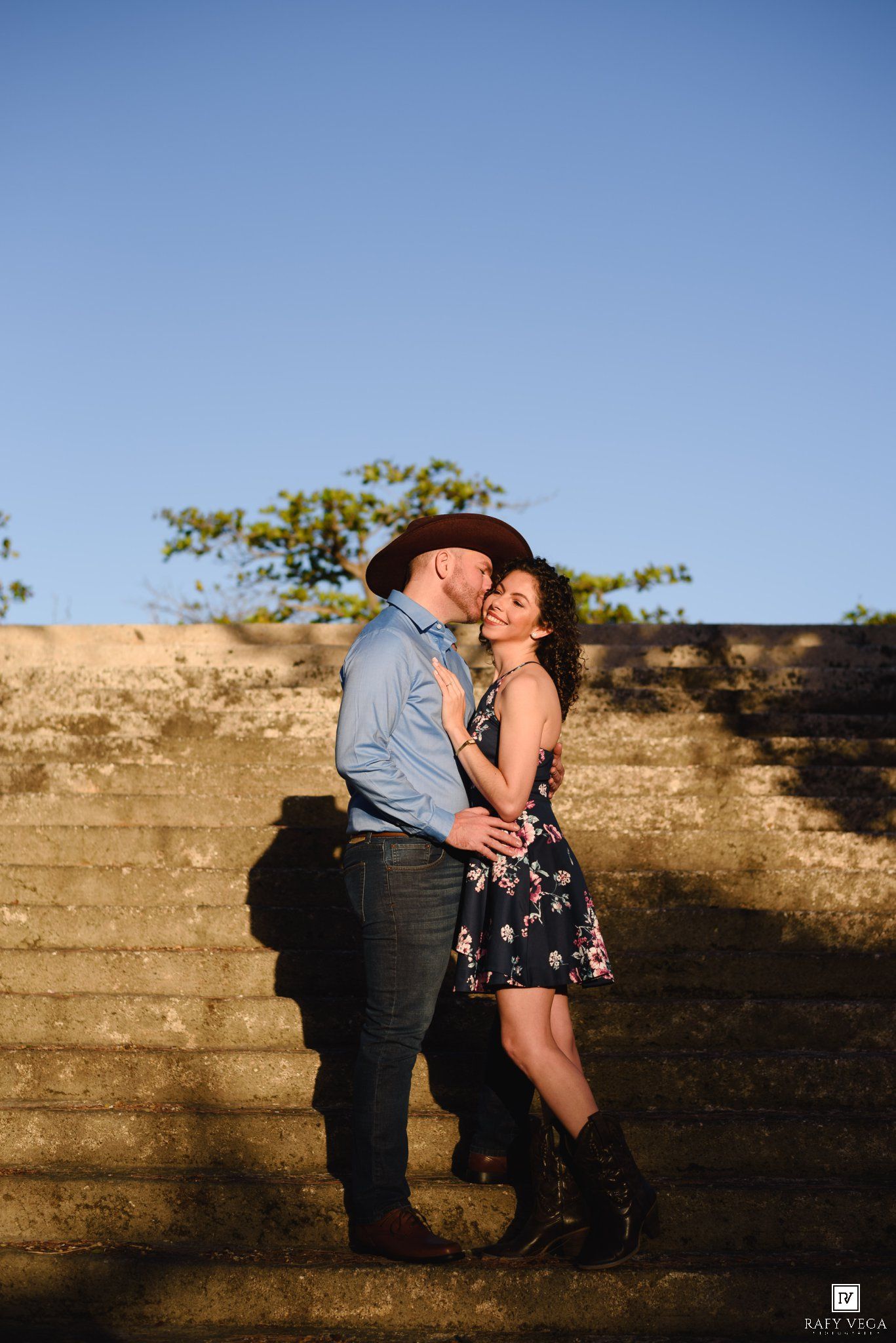 Couple embracing on stone steps at golden hour, man in cowboy hat and woman in floral dress with boots.