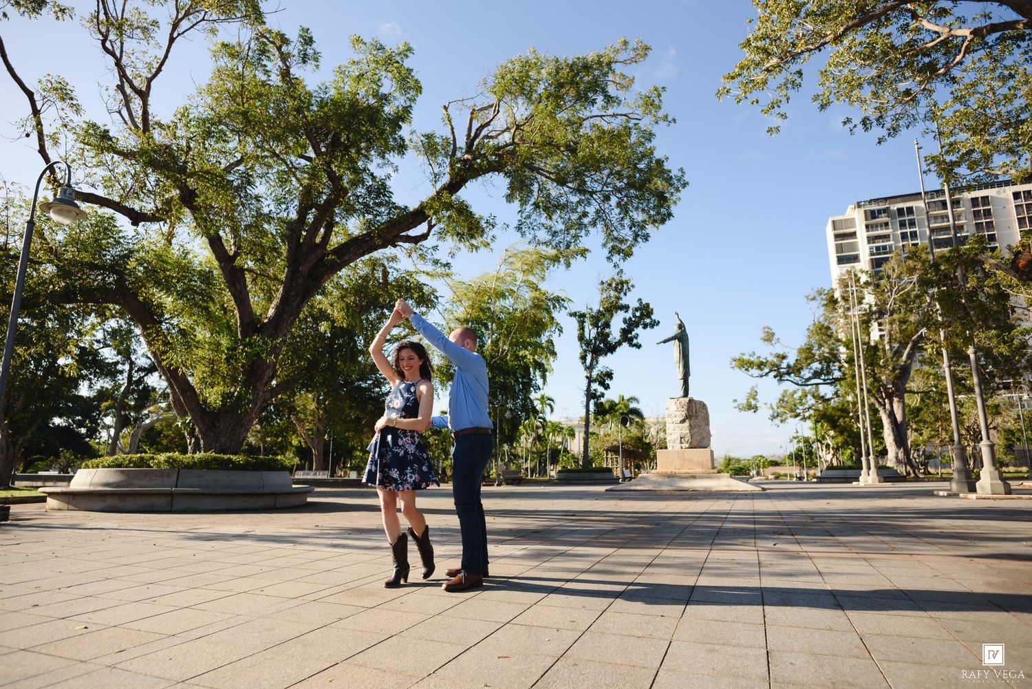 Couple dancing joyfully in an open plaza surrounded by trees and a statue on a bright sunny day.