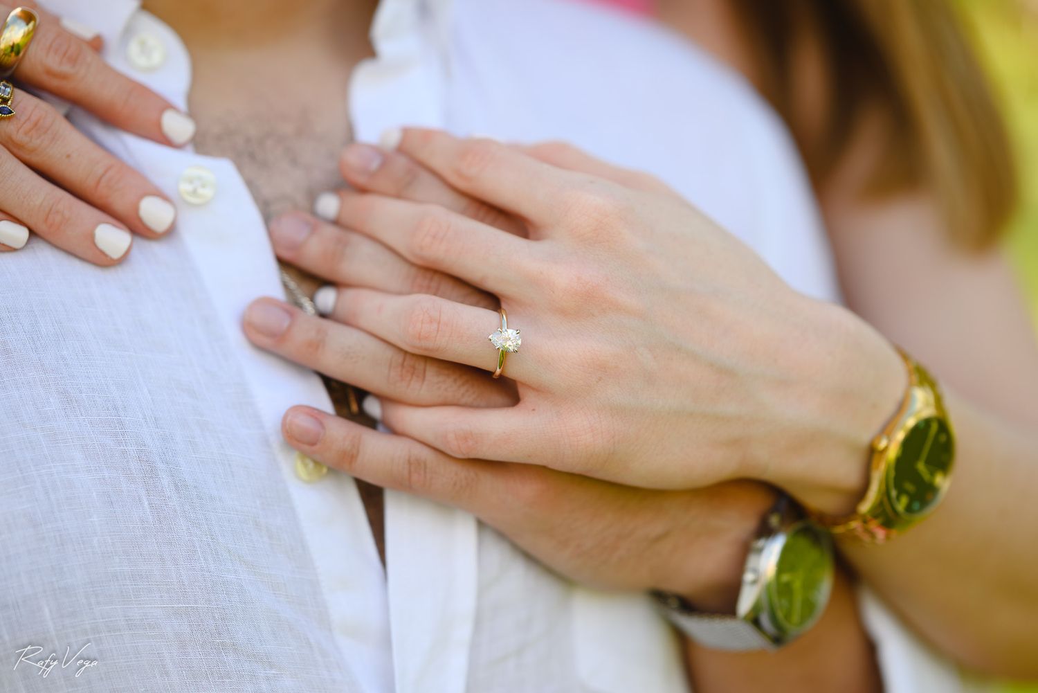 Close-up of couple embracing, showing engagement ring and gold bracelet on woman's hand resting on man's white shirt.