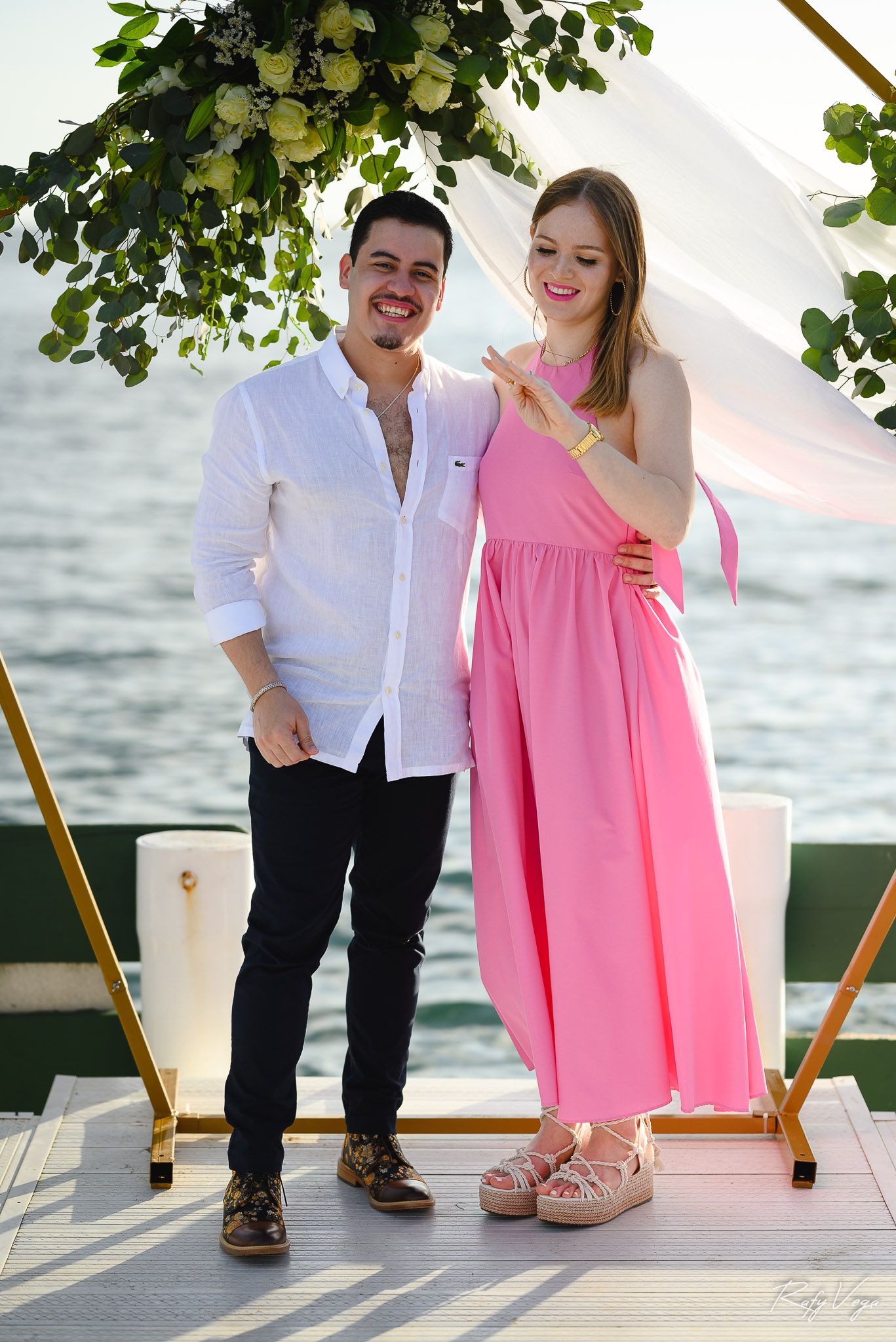 Couple posing together on a waterfront platform, man in white shirt and woman in pink maxi dress under floral arch.