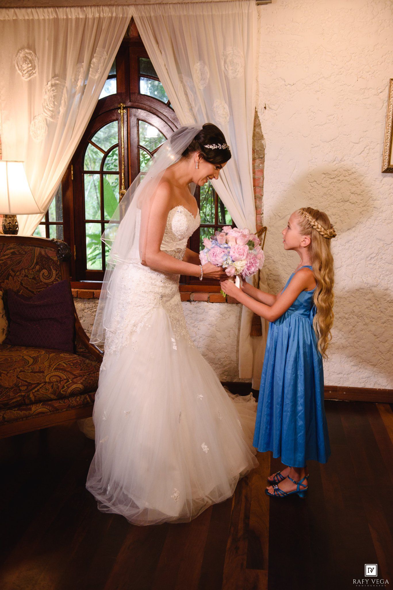 Bride in white gown receives pink bouquet from young flower girl in blue dress indoors.