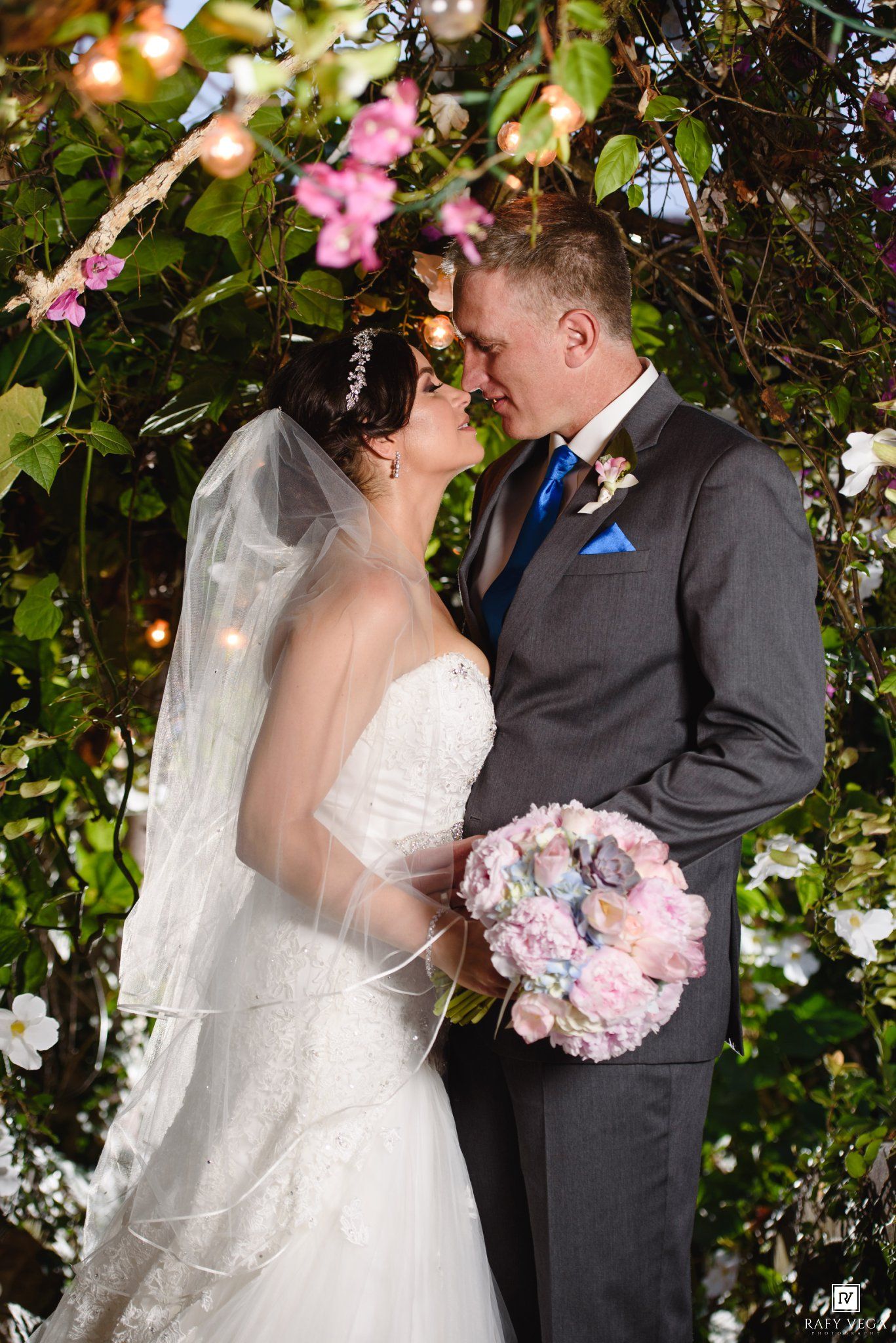 Bride and groom touch foreheads tenderly under pink floral arch, bride holding pastel bouquet.
