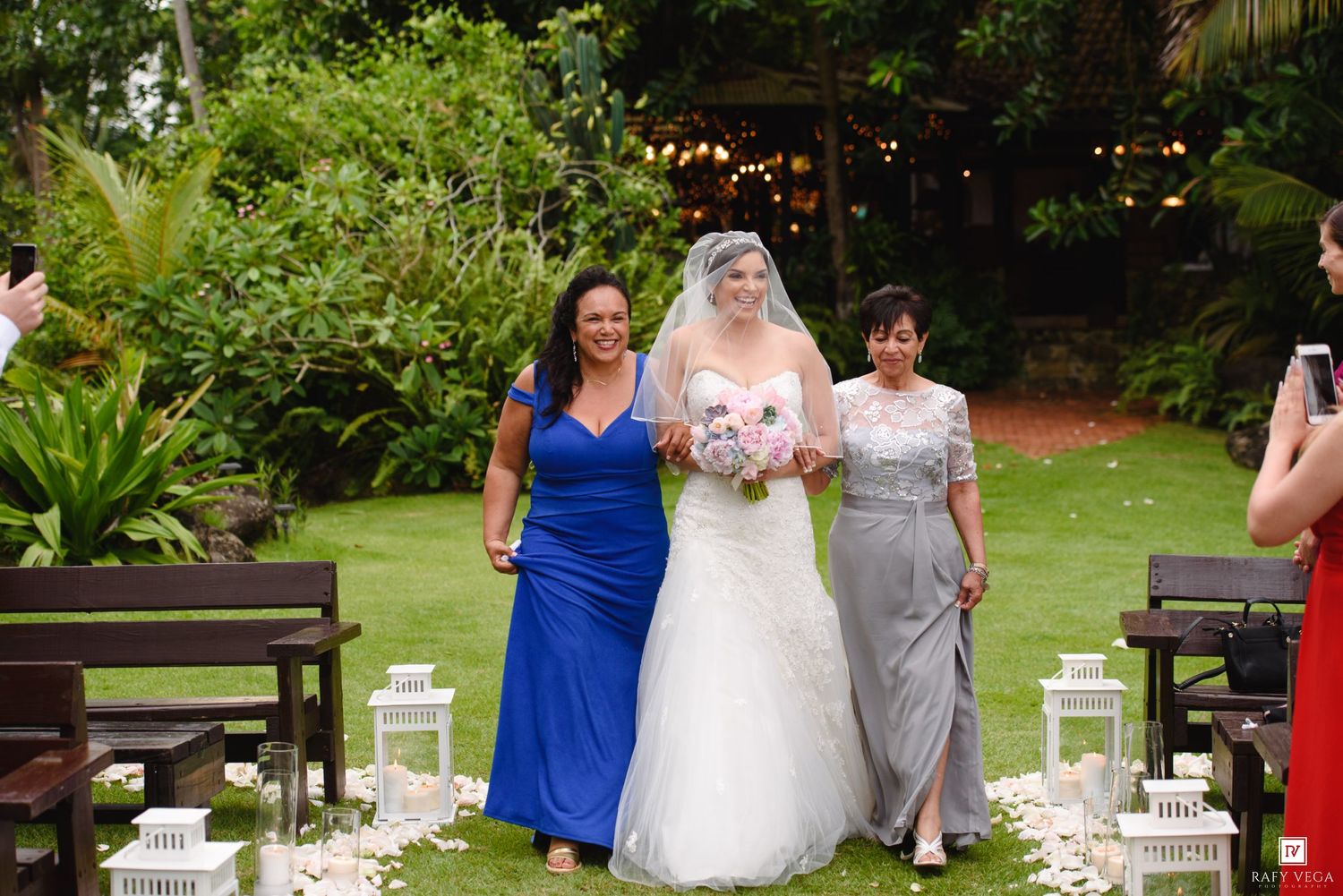 Bride walks down outdoor aisle flanked by two women in blue and grey dresses, surrounded by lush greenery.