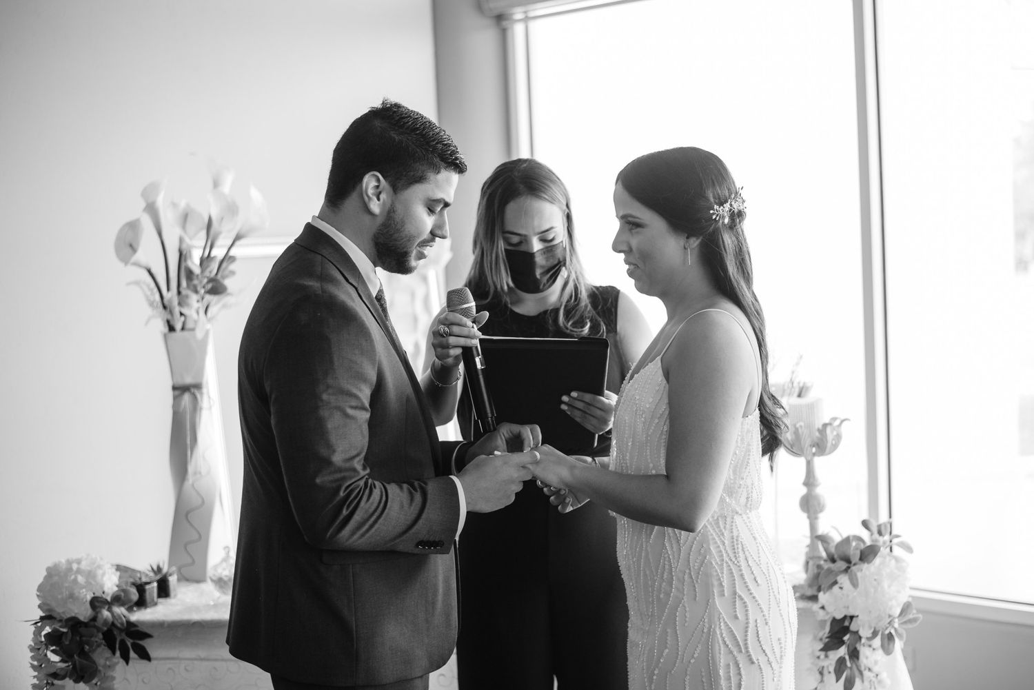 Black and white wedding ceremony photo of couple exchanging rings with officiant wearing mask indoors.