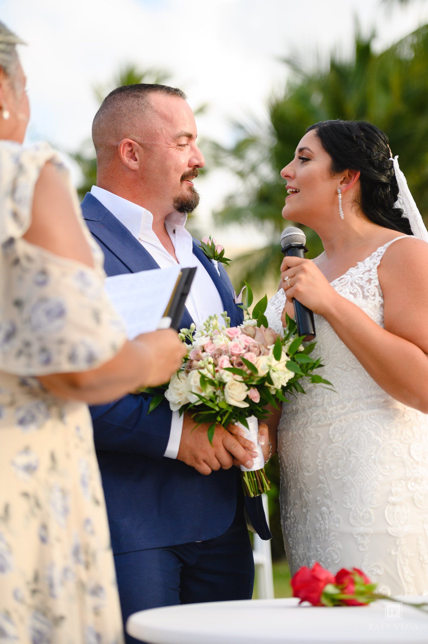 Bride reads vows to groom at outdoor wedding ceremony, holding pink and white bouquet, officiant visible in foreground.