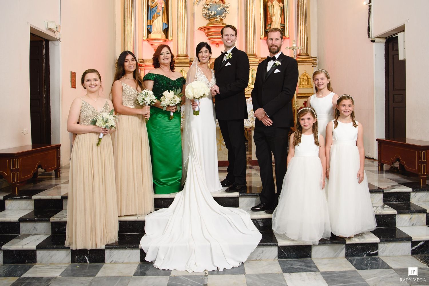 Wedding party poses in church with bride in white gown, bridesmaids in green and gold dresses, groom in black suit, and flower girls.