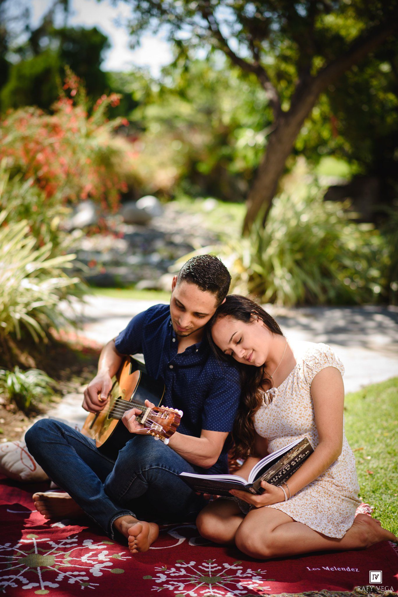 Couple sitting on red blanket in garden, man playing acoustic guitar while woman leans on him reading a book.