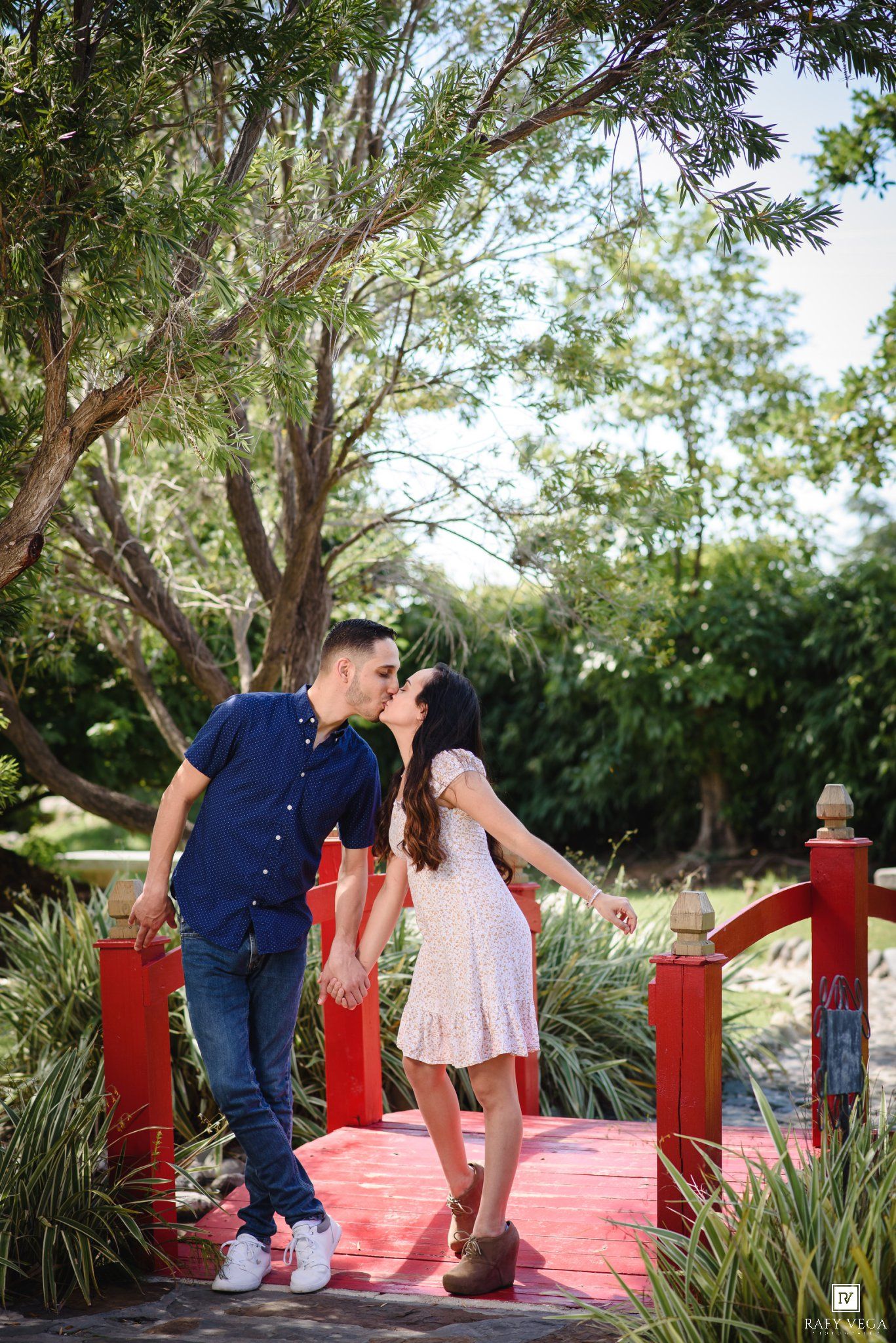 Couple sharing a romantic kiss on a red bridge surrounded by lush green trees in a garden setting.
