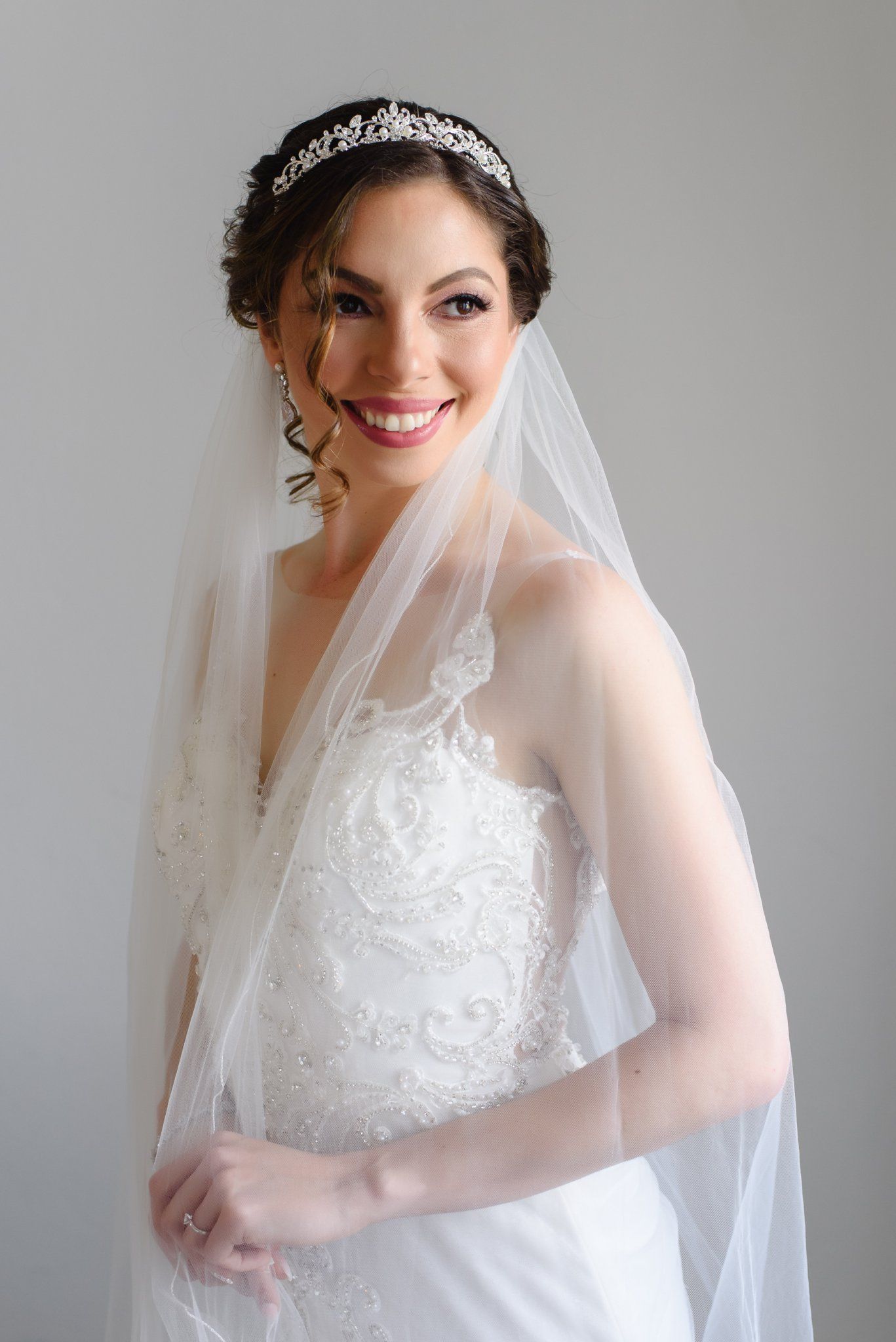 Smiling bride in lace wedding gown, tiara, and veil poses elegantly against a light gray background.