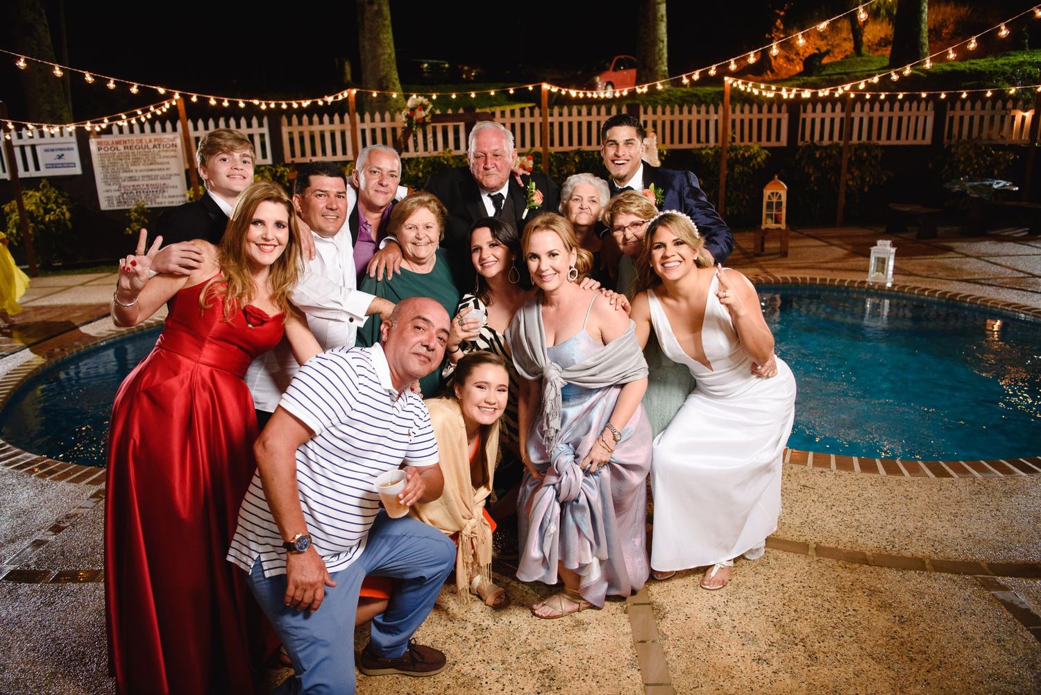 Large family group posing for selfie by pool at outdoor wedding reception with string lights at night.