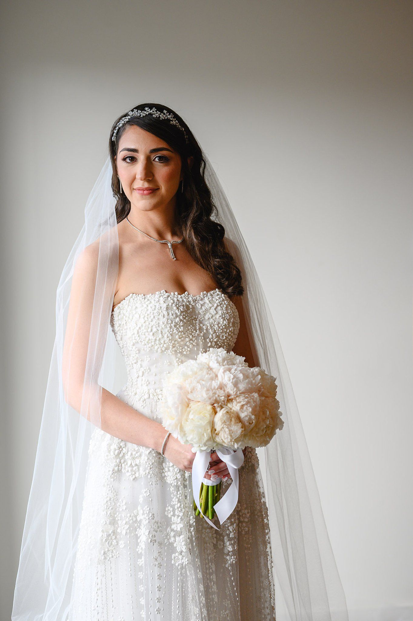 Bride in strapless pearl-embellished gown with veil and tiara holds white peony bouquet.