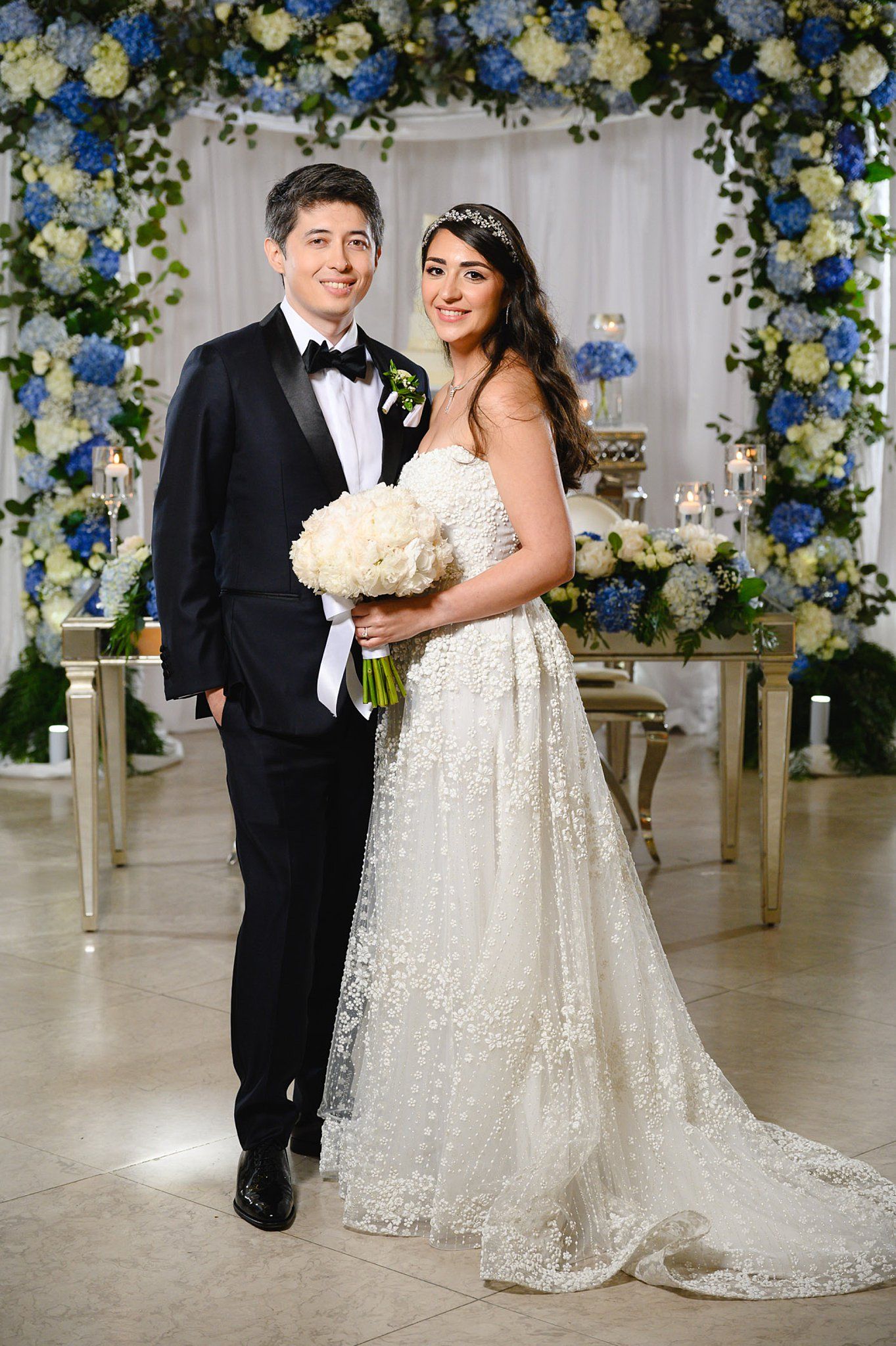 Bride in lace gown and groom in tuxedo pose together before blue and white floral arch at elegant wedding ceremony.