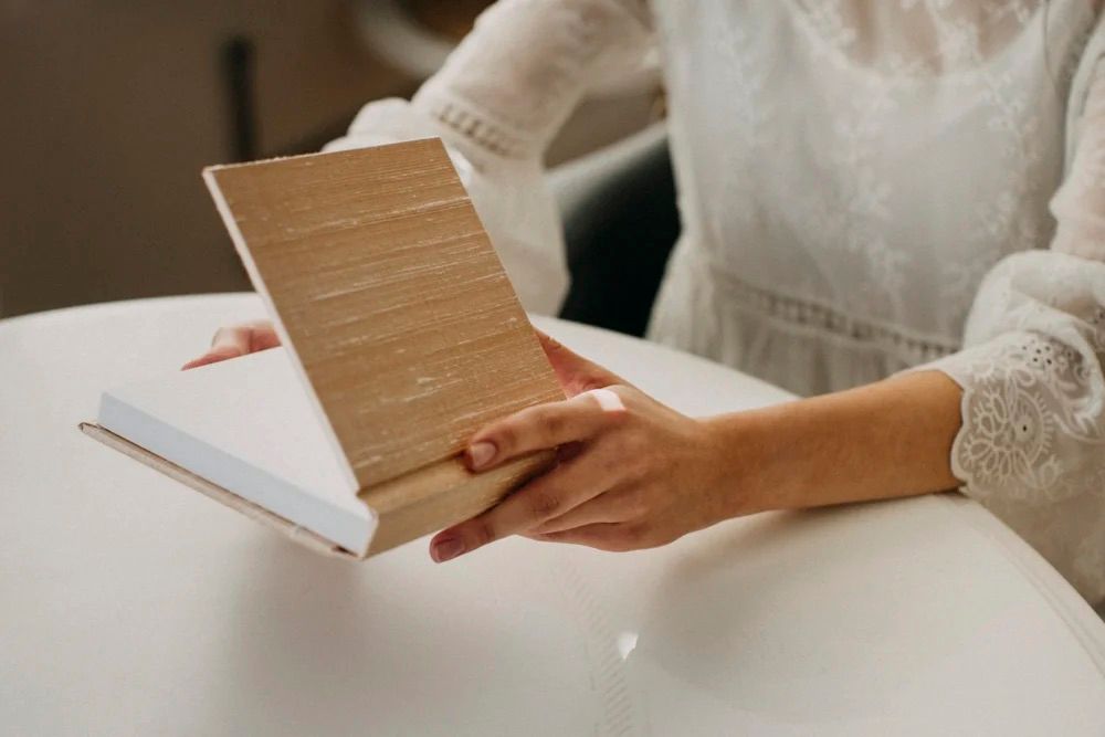 Woman in white lace dress holding a wooden hardcover book at a white table.