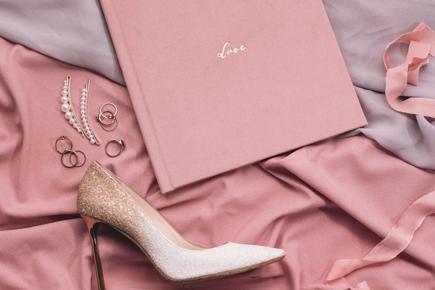 Pink satin fabric with glitter heels, pearl earrings, gold rings, ribbon, and a pink Love journal flatlay.