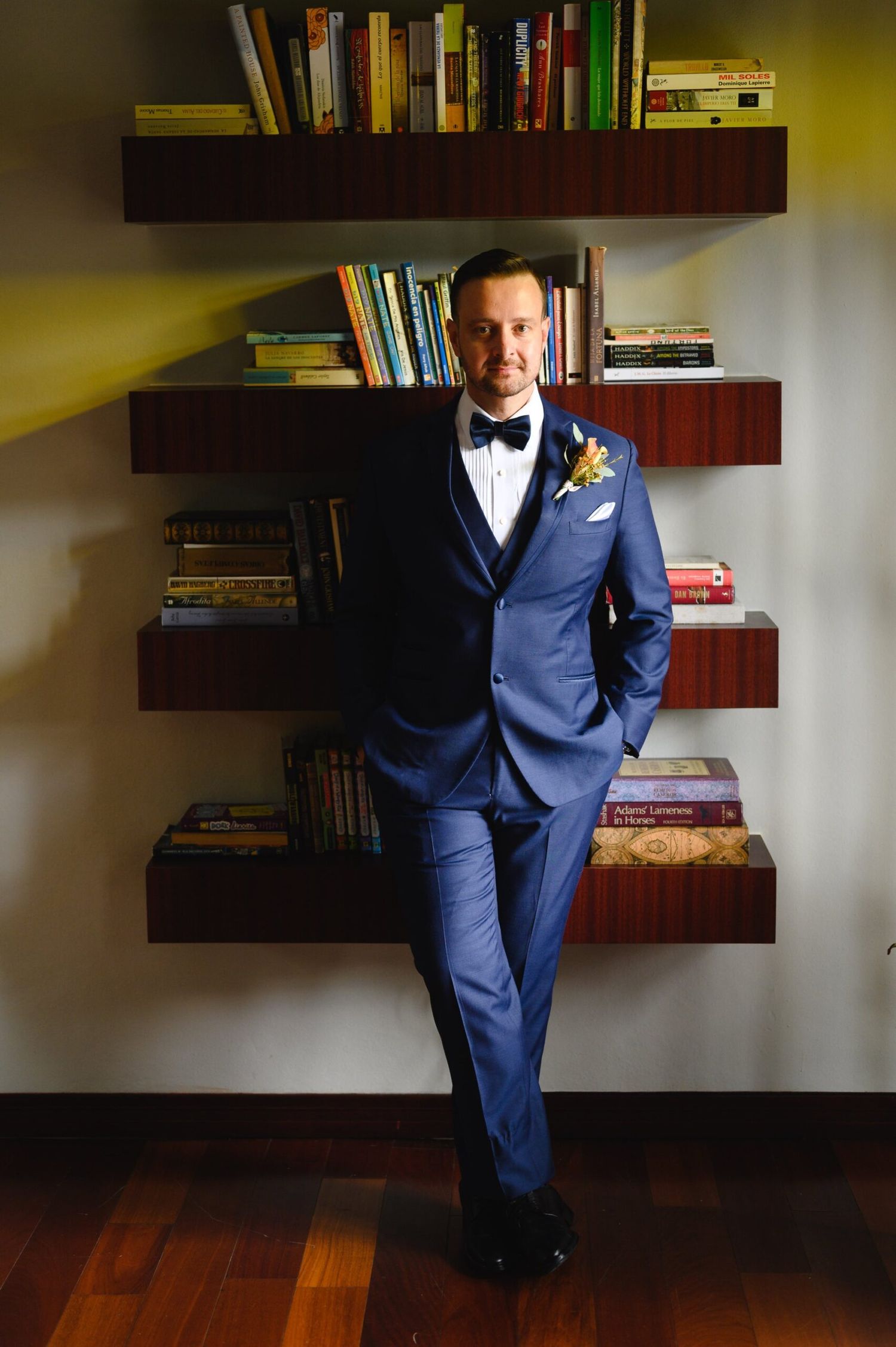 Groom in blue tuxedo with bow tie and boutonniere poses confidently in front of floating bookshelves.