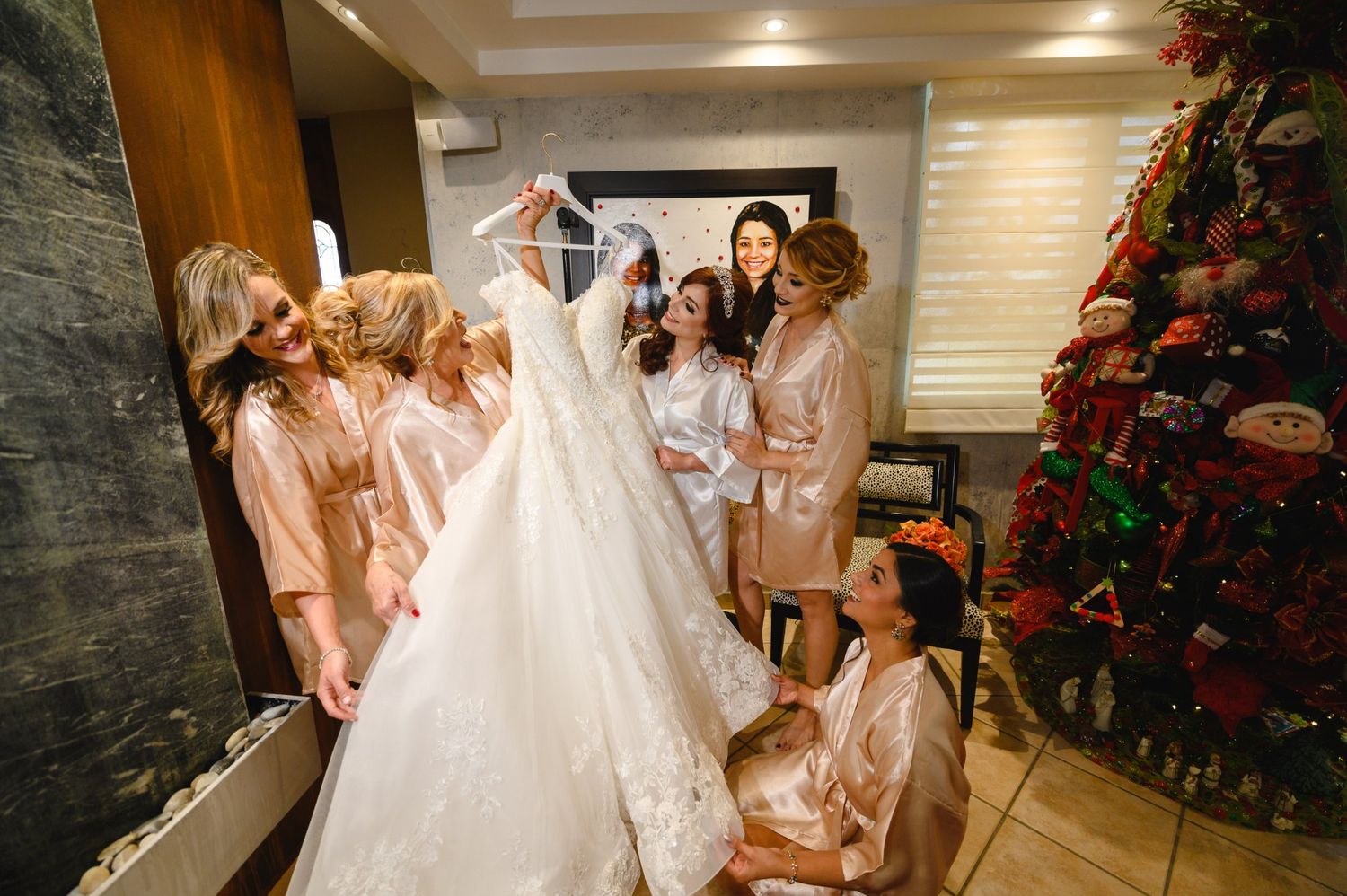 Bride holding wedding gown with bridesmaids in robes gathered around in a decorated room.