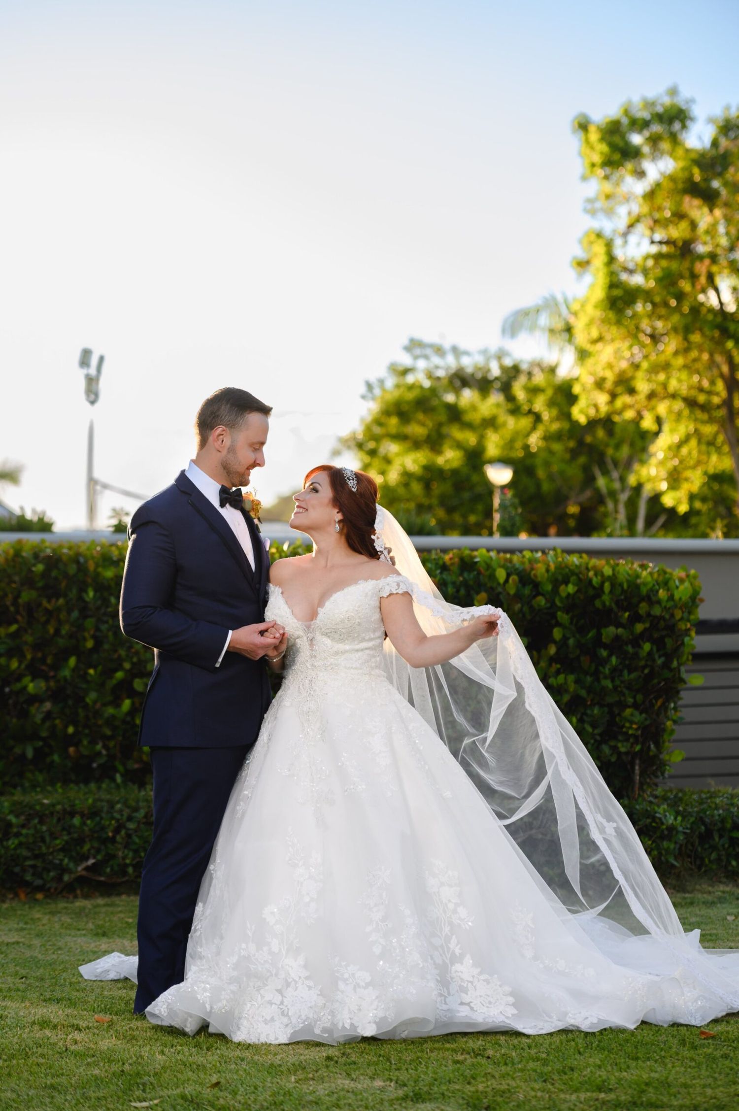 Bride in lace ball gown and veil shares romantic moment with groom in navy tuxedo on green lawn.