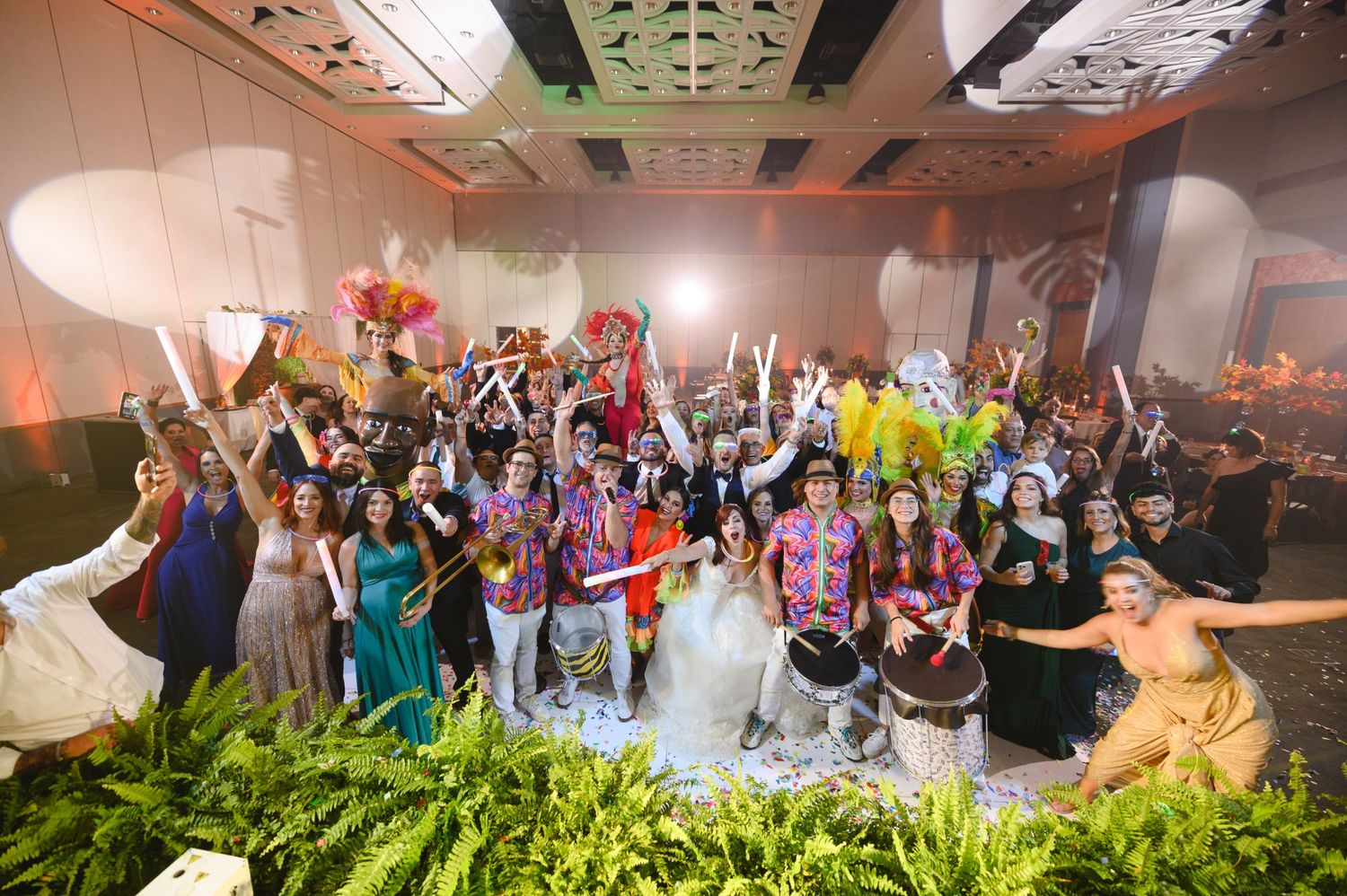 Large wedding party celebrating on dance floor with colorful outfits, raised arms, and festive decorations in elegant venue.
