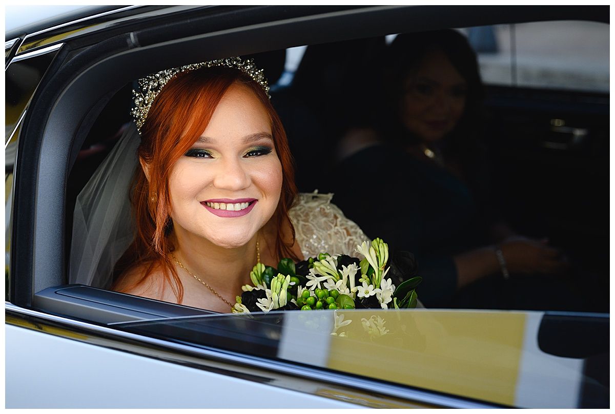 Smiling bride with red hair and tiara holding bouquet inside a car on her wedding day.