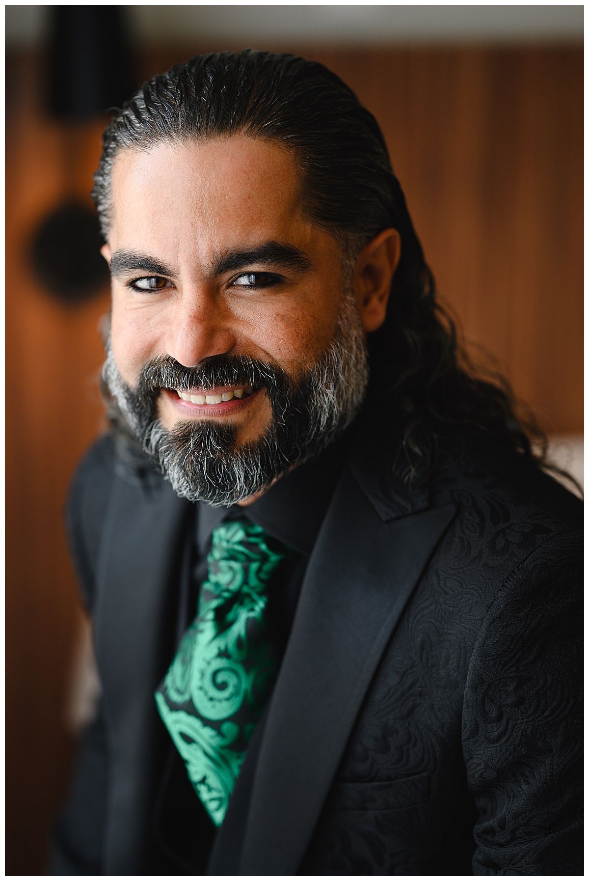Smiling man with long dark hair and beard wearing a black paisley suit with a green tie.
