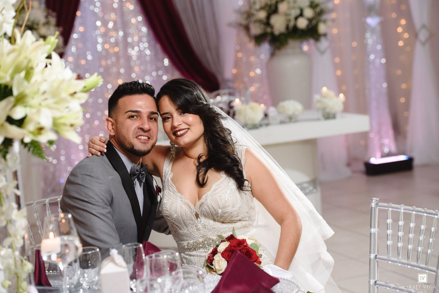 A bride and groom smile together at their elegant wedding reception, surrounded by floral arrangements and twinkling lights.