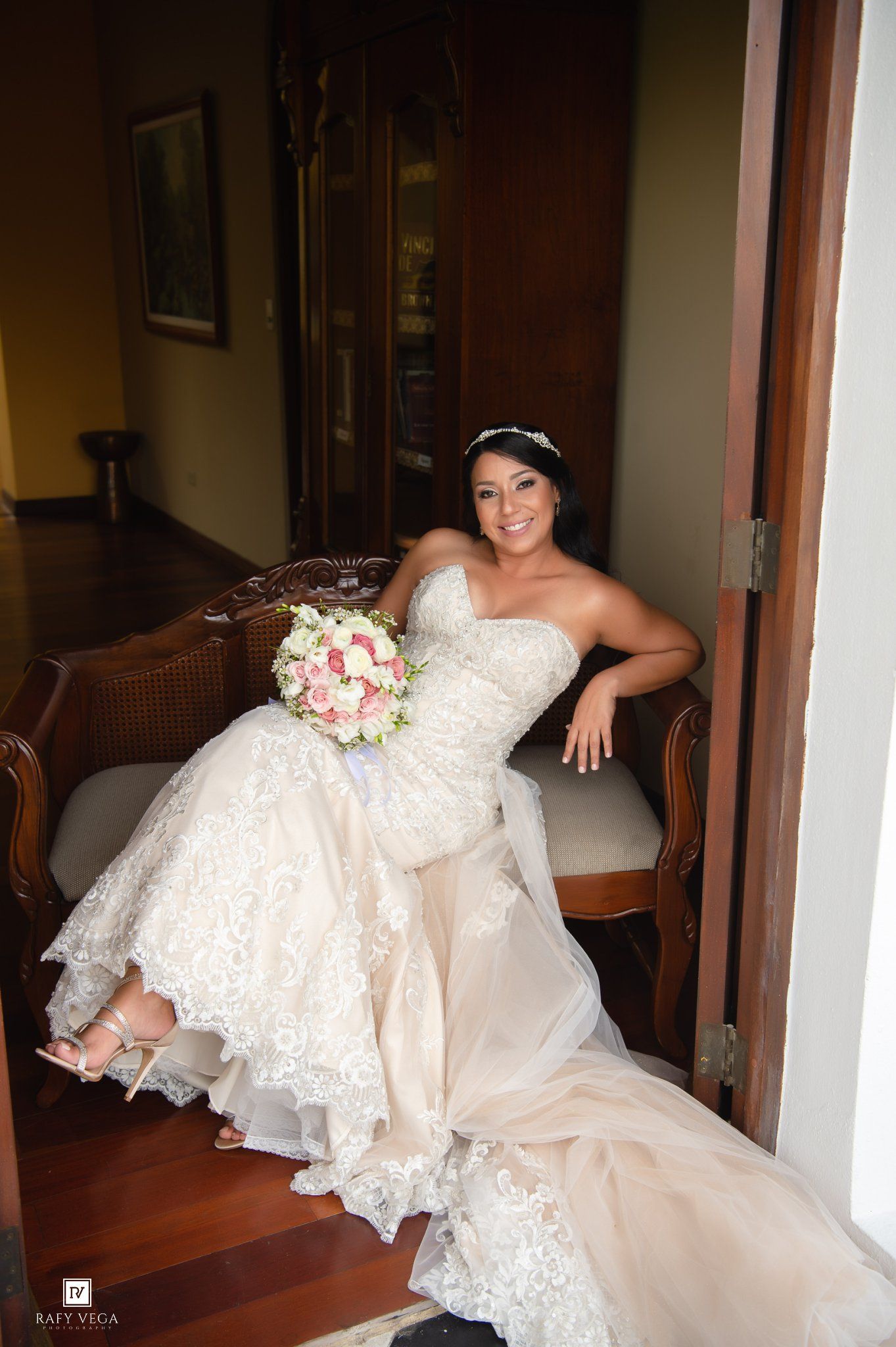Smiling bride in strapless lace wedding gown holding pink bouquet, seated elegantly on vintage bench in warmly lit hallway.