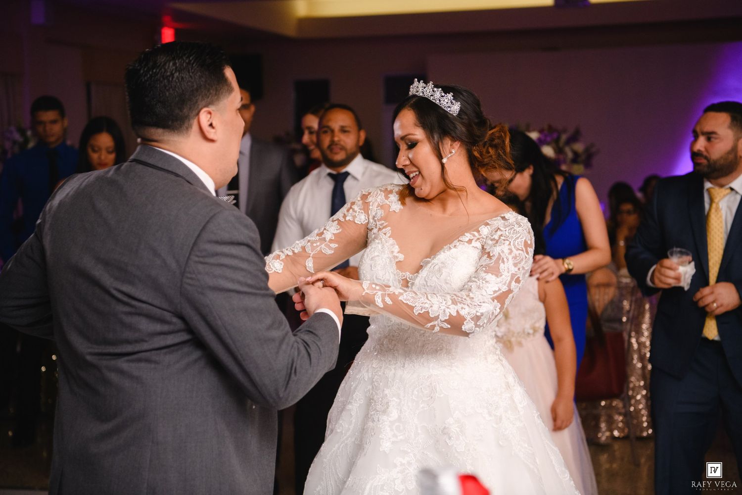 Bride in lace gown and groom share first dance surrounded by guests at elegant wedding reception.