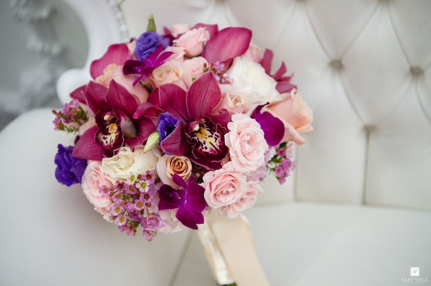 Bridal bouquet with purple orchids, pink roses, and violet blooms arranged on a white tufted chair.