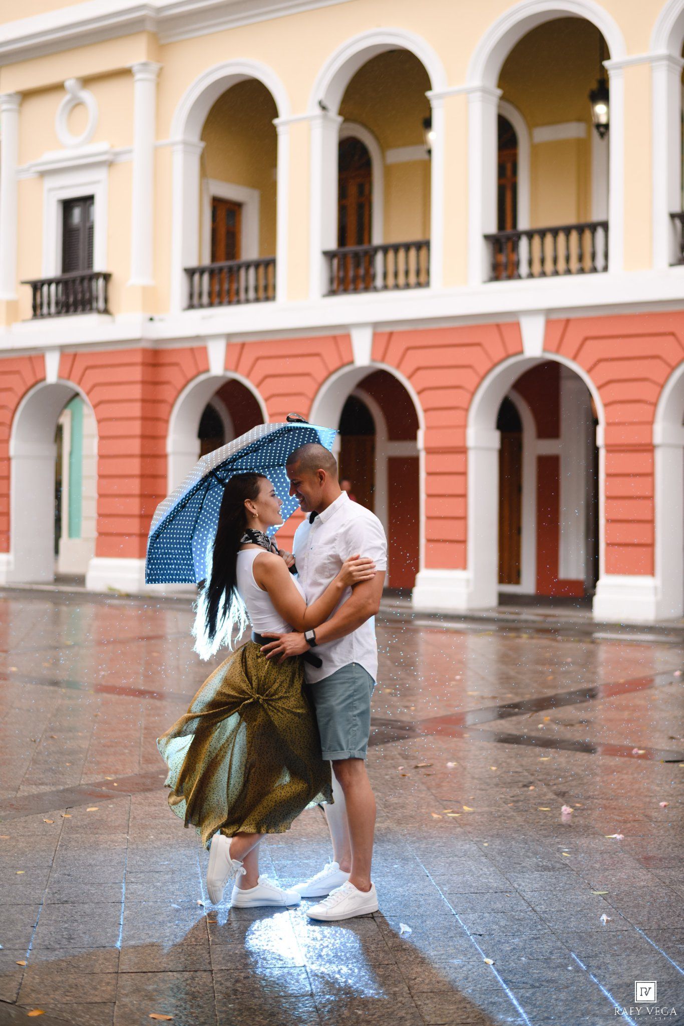 Couple sharing a romantic kiss under a blue umbrella in the rain outside a colorful colonial building.