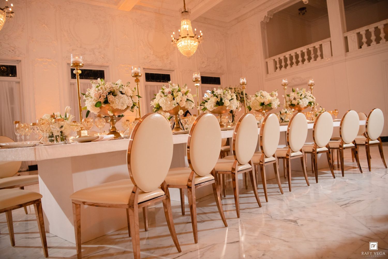 Elegant wedding reception with gold chairs, long white tables, and floral centerpieces in a grand ballroom.