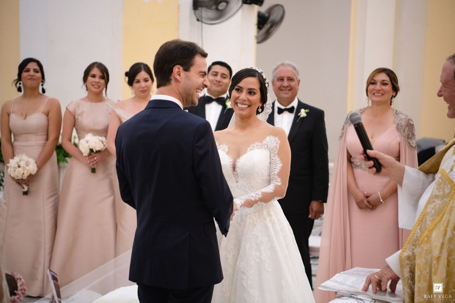 Bride and groom exchange vows at church altar surrounded by bridesmaids in pink dresses and groomsmen in tuxedos.