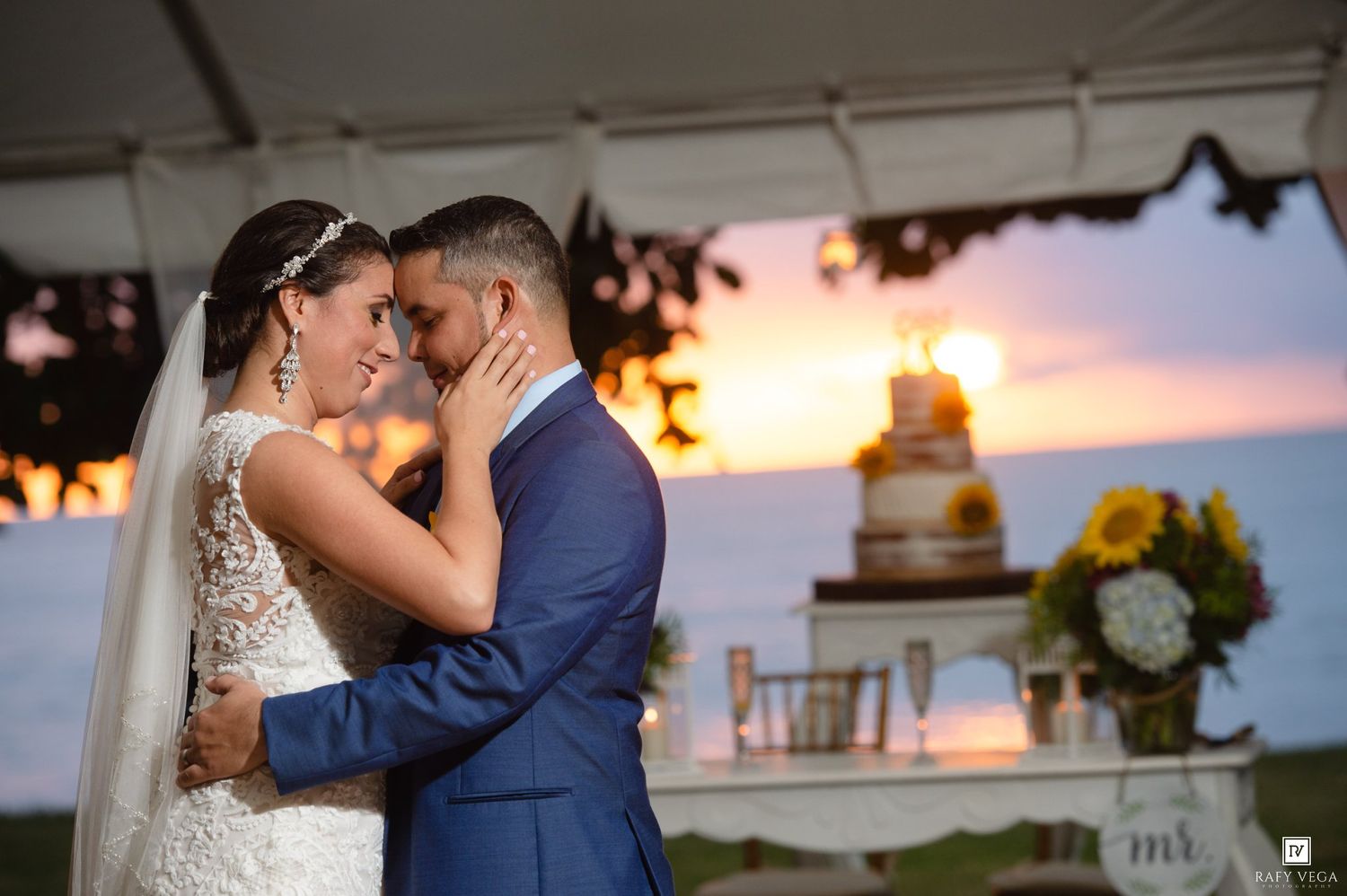 Bride and groom share a romantic sunset embrace at outdoor wedding reception with tiered cake and sunflowers in background.