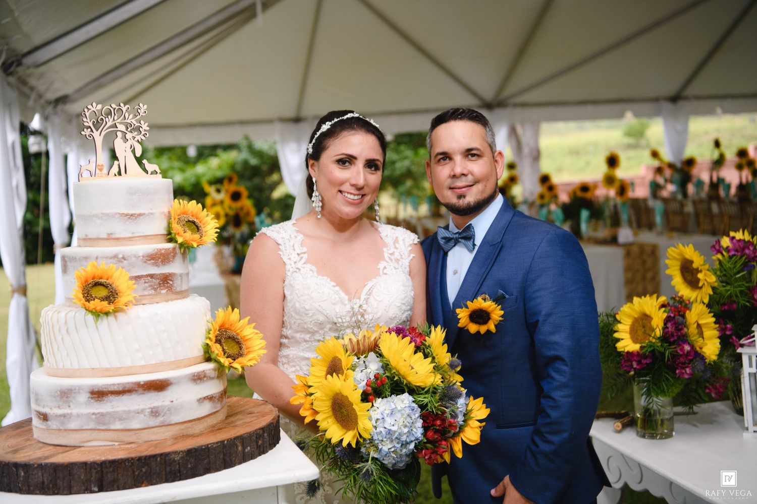 Smiling bride and groom posing beside a rustic tiered wedding cake decorated with sunflowers at their tented reception.