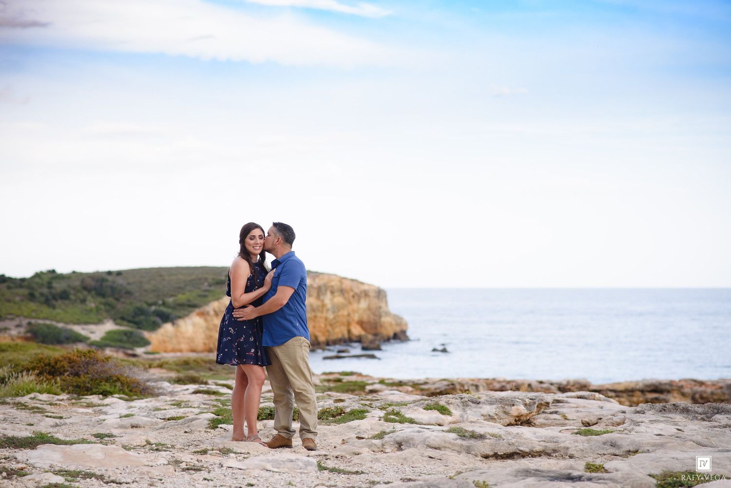 Couple embracing romantically on rocky coastal cliffs with ocean and headlands in the background.