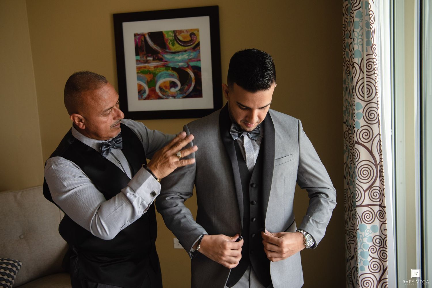 Two groomsmen helping each other button up gray suits before the wedding ceremony.
