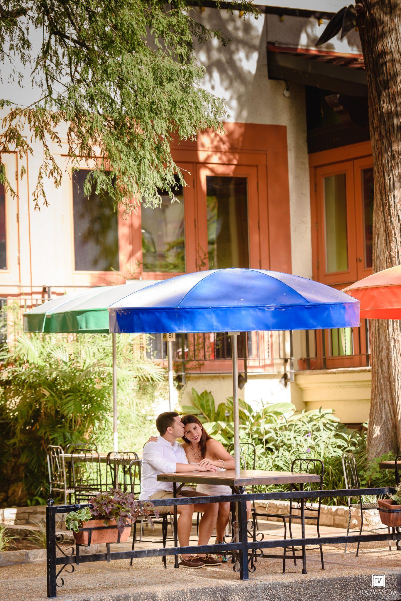 Couple sitting together under a blue patio umbrella at an outdoor cafe surrounded by lush greenery.