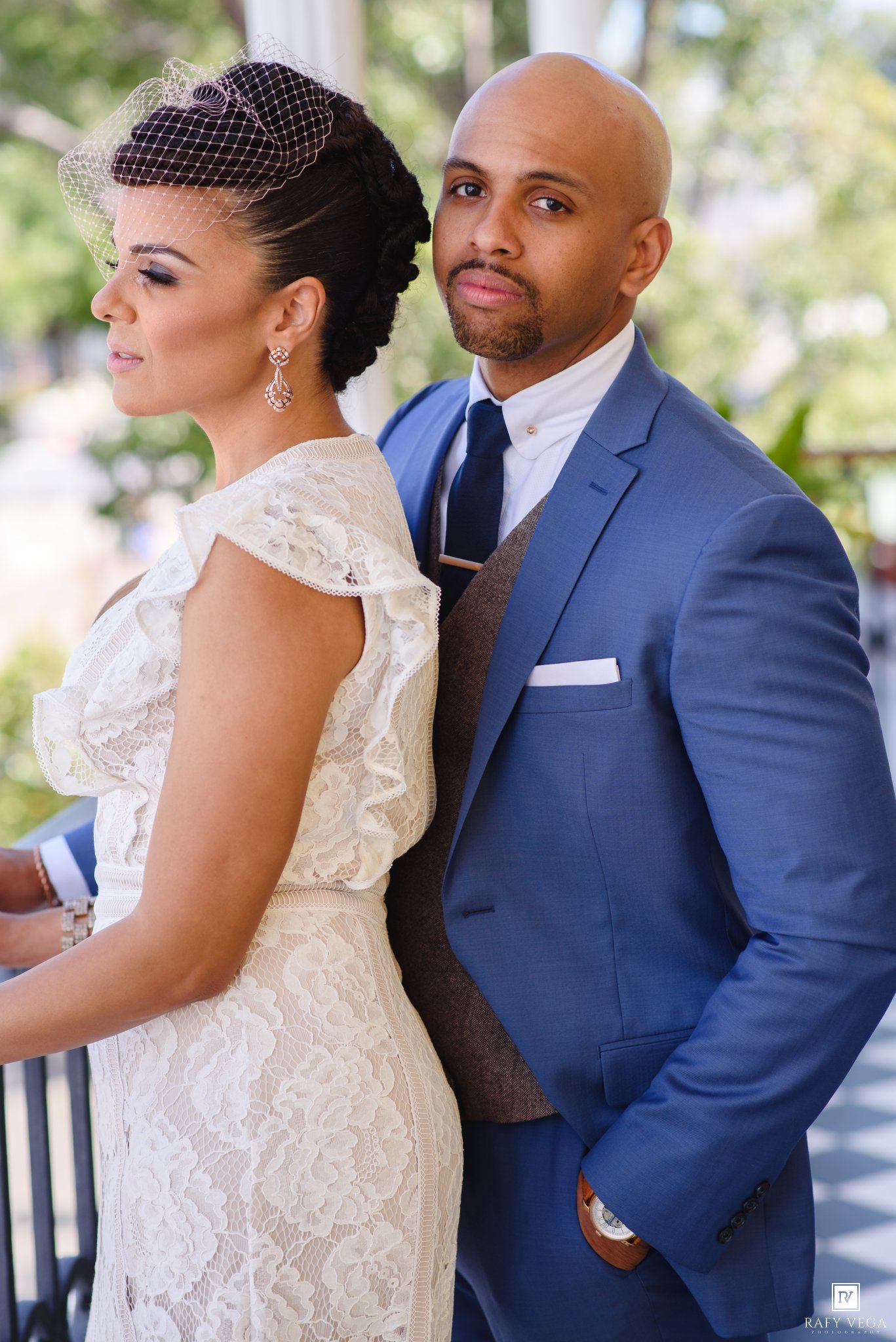 Elegant bride in lace dress and groom in blue suit posing back-to-back at a wedding ceremony.