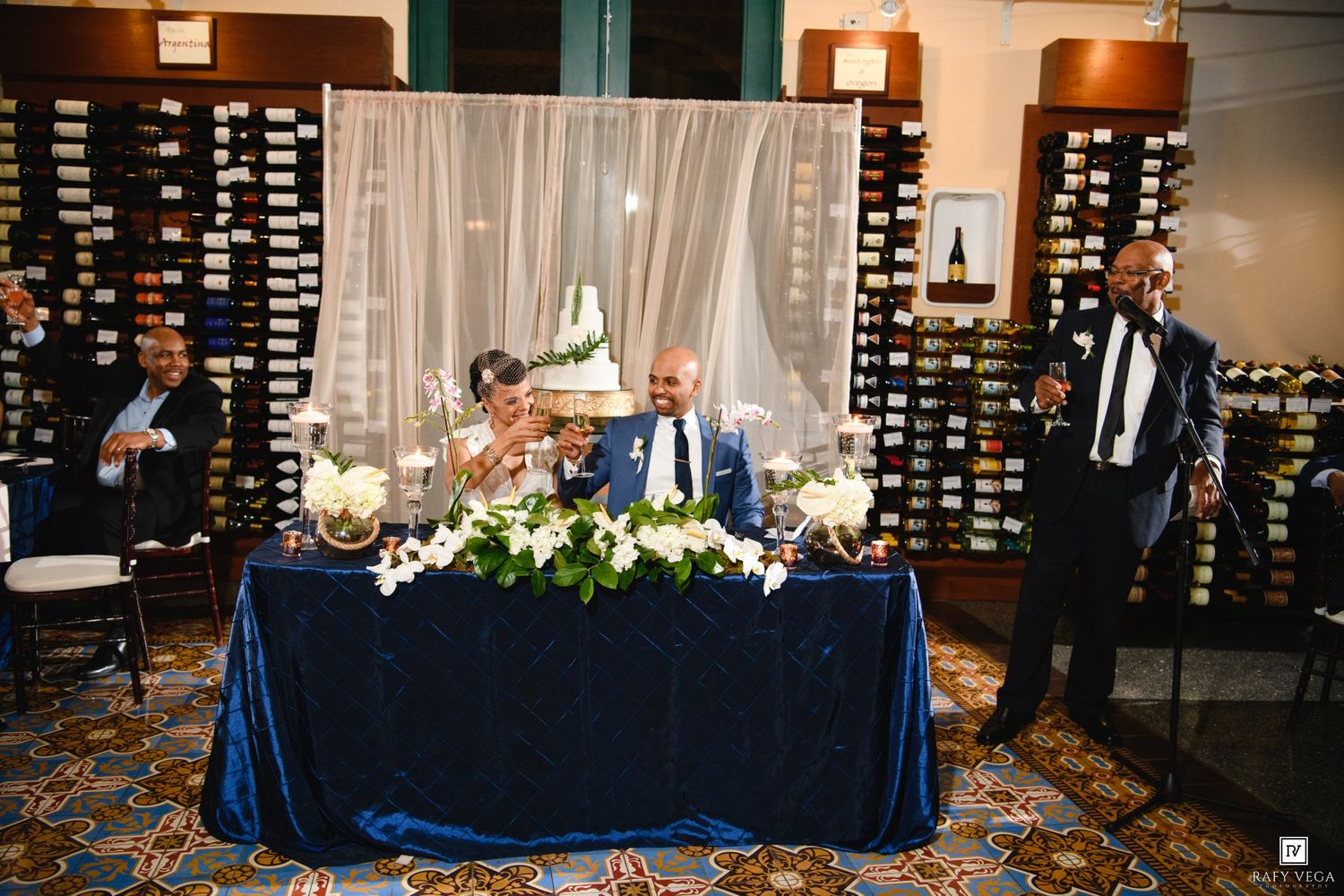 Couple seated at elegant sweetheart table with blue velvet linen, white floral centerpieces, and guests celebrating at wedding reception.
