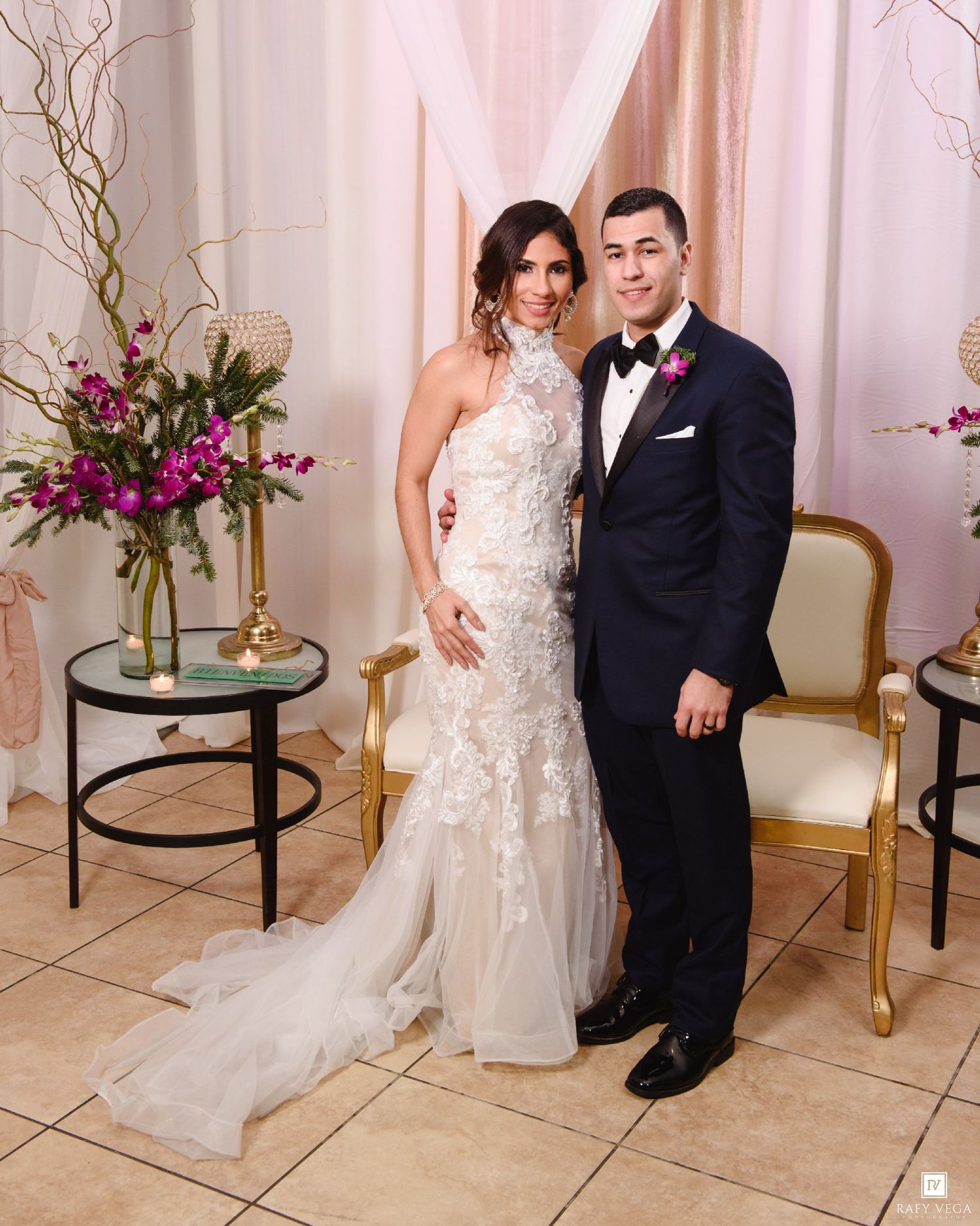 Bride in lace mermaid gown and groom in navy tuxedo pose together at elegant wedding venue with floral decor.