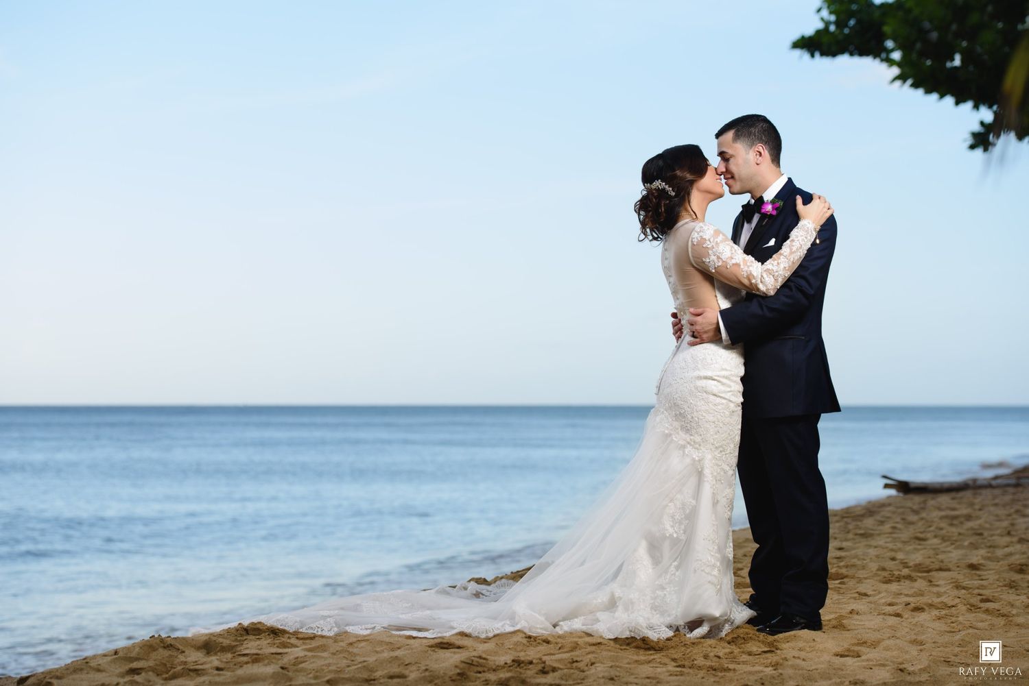Bride and groom embrace romantically on sandy beach at dusk with calm ocean backdrop.