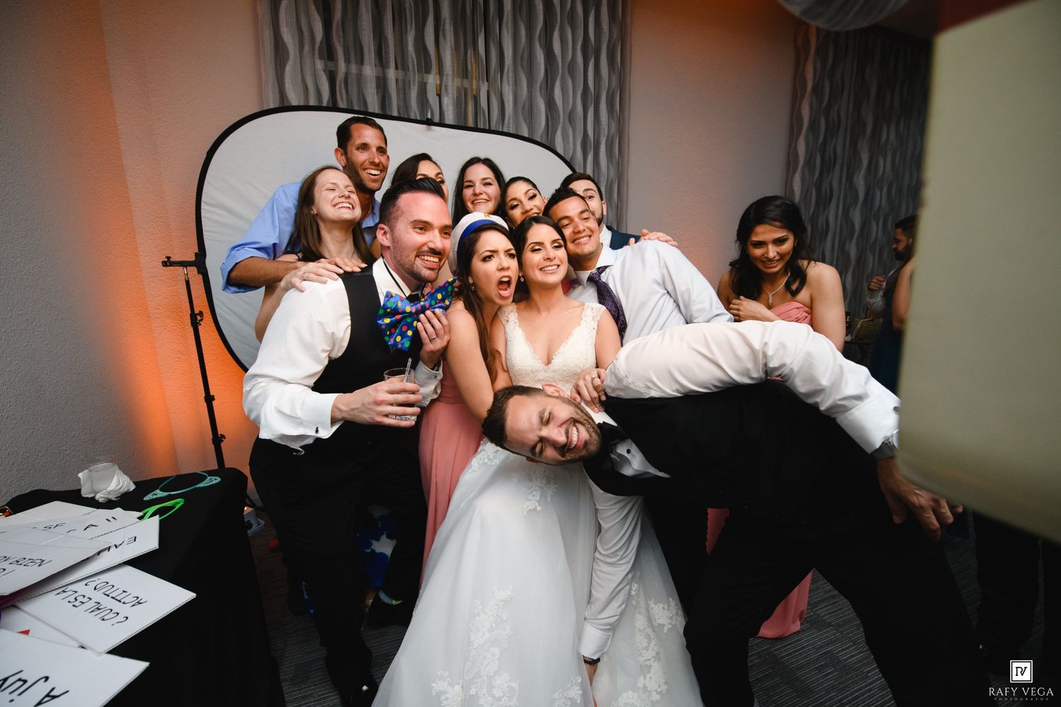 Wedding party group photo with bride and groom surrounded by smiling guests at a reception venue.