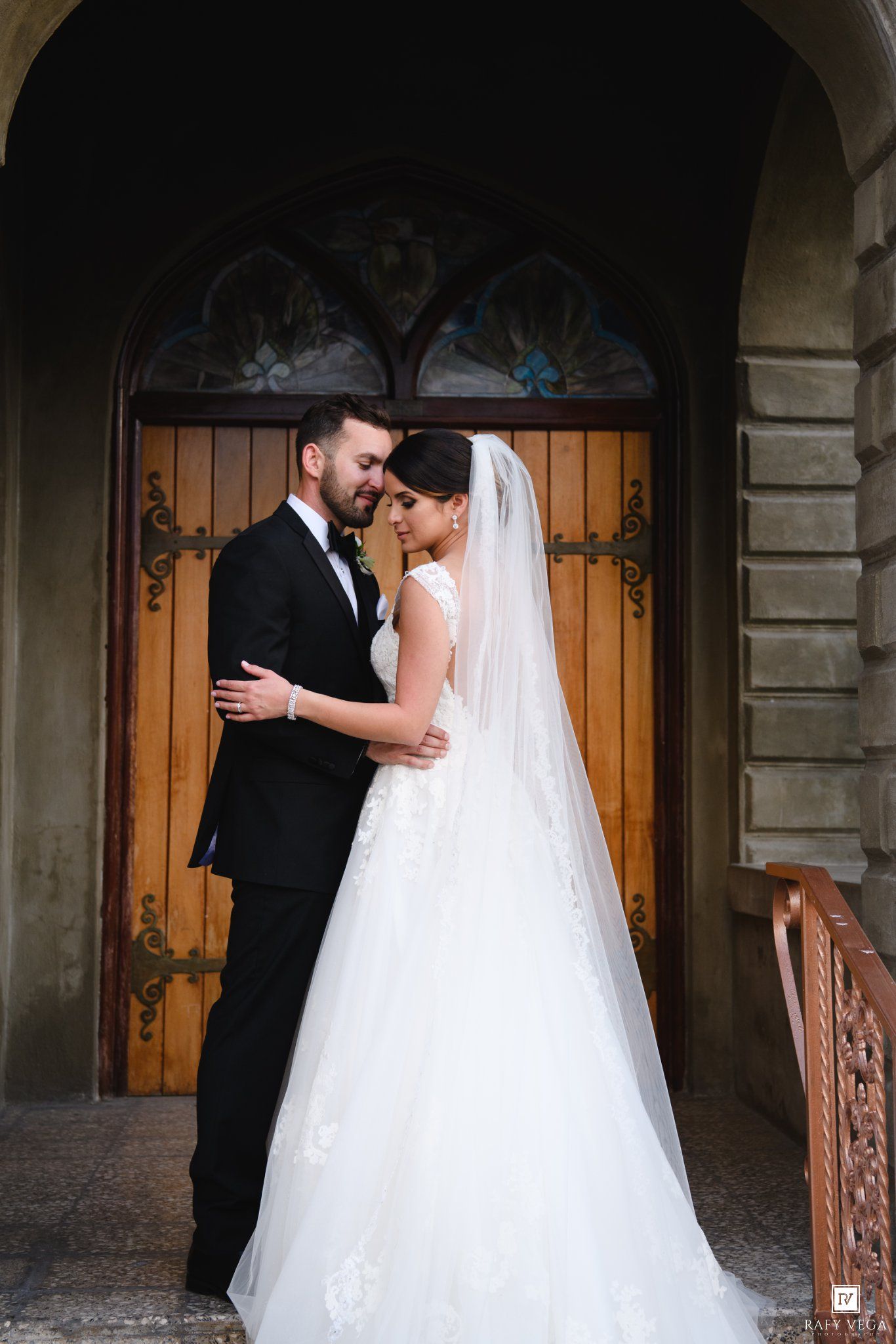 Bride in white gown and veil with groom in black tux embracing before ornate wooden church doors.